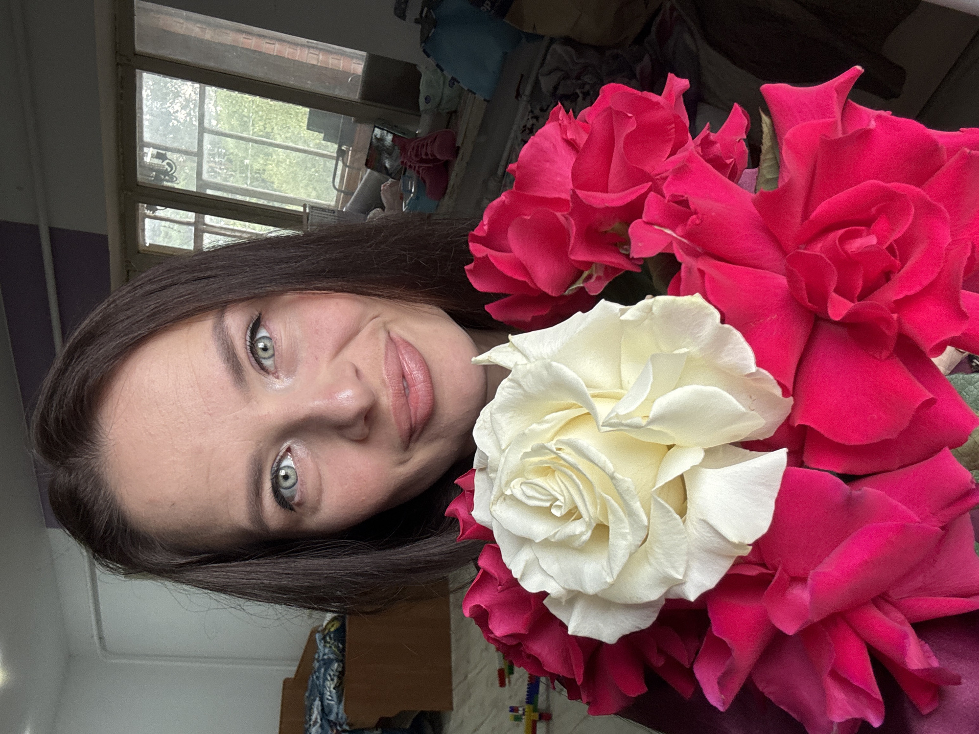 A woman with brown hair and blue-green eyes is holding a bouquet of roses, with a large white rose in the center surrounded by several bright pink roses. She is smiling slightly and looking towards the camera. The background appears to be an indoor setting, possibly a room in a house or apartment, with a window visible in the upper right corner showing trees and daylight. The room appears somewhat cluttered with furniture and other items. The location of Rusalevka, Russia, is not discernable from the visual information in the image.