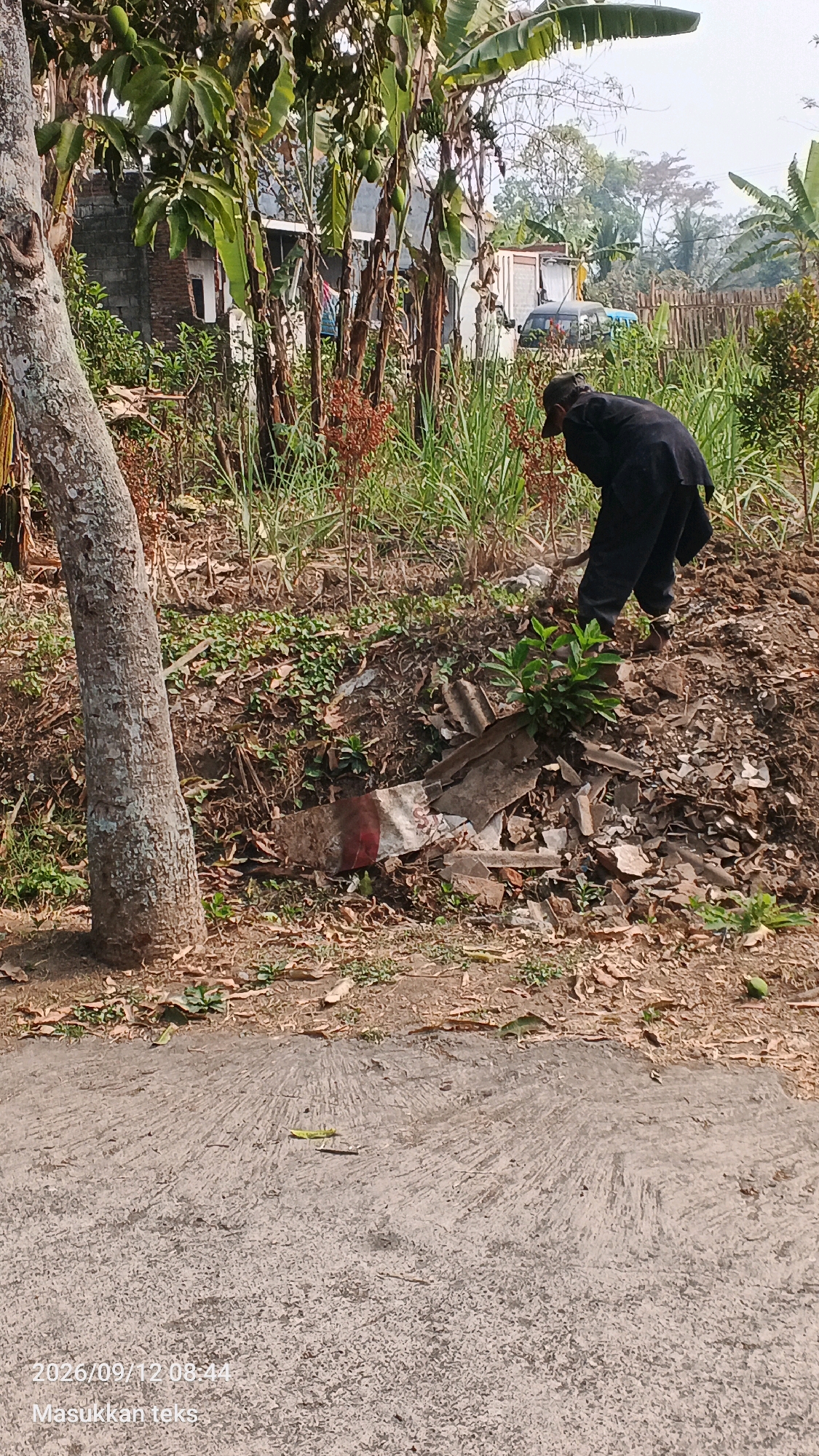 In Jeru, Indonesia, a person wearing a dark outfit is bent over and appears to be working on a pile of dirt and debris. The area is overgrown with vegetation, including tall grasses and trees. In the background, a brick building is visible, along with a white shipping container and parked vehicles. The ground in the foreground is a paved surface with a textured pattern. A timestamp in the lower left corner reads "2026/09/12 08:44" and the text "Masukkan teks" is also present.