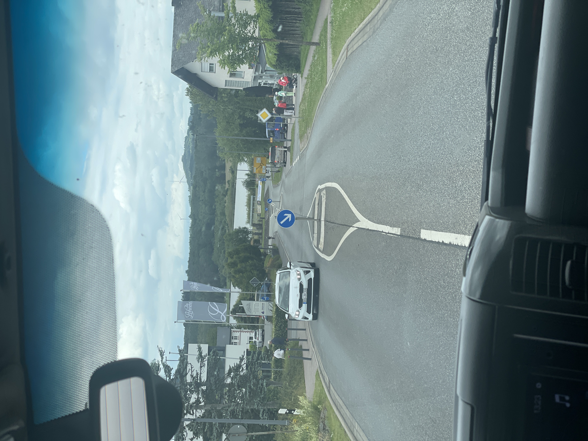 This image captures a scene on a street in Ulmen, Germany, likely on a cloudy day given the overcast sky. The perspective is from inside a vehicle, looking forward through the windshield. The road ahead shows lane markings and a directional arrow sign indicating a turn.
Several vehicles are present on the road. A white Kia hatchback with license plate "DAU-SH 168" is in the foreground, traveling in the same direction as the viewer. Further down the road, a silver minivan and a blue panel van are visible. On the right side of the road, near a building with a dark fence, a group of people are gathered. Among them are individuals in casual attire, and two individuals appear to be dressed in bright red uniforms, one of whom is on what looks like a mobility scooter or a small motorized cart.
To the left of the road, there are trees, a building with multiple windows, and flags displaying the logo "Lohners". In the background, a body of water can be seen, with a treed hillside and wind turbines on the horizon. The overall environment suggests a semi-urban or suburban setting.