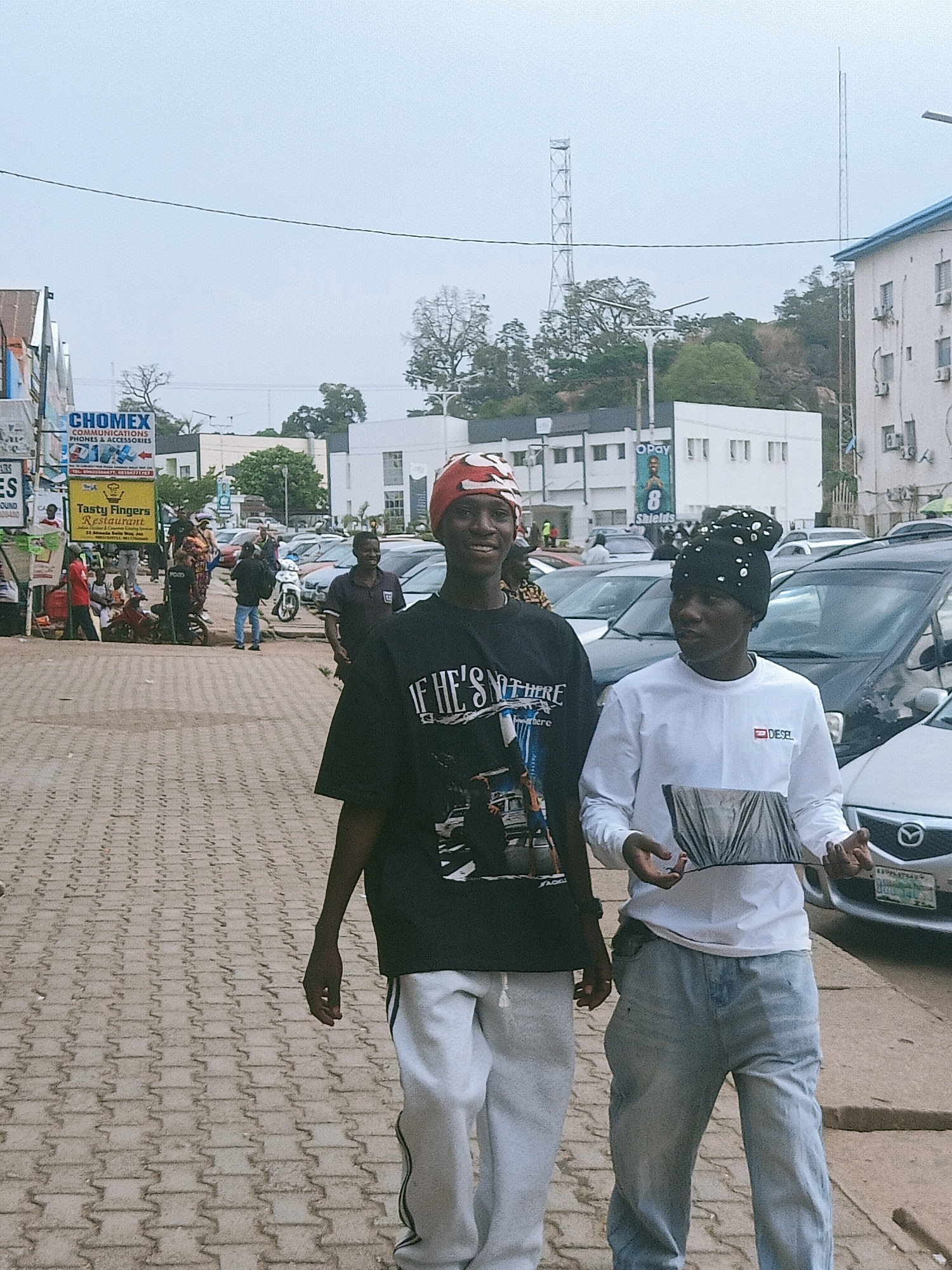 The image shows two young men walking on a paved sidewalk in a busy urban environment. The man on the left wears a black t-shirt, white sweatpants, and a red head covering. The man on the right wears a white long-sleeved shirt, blue jeans, and a black knit cap with white adornments. Cars are parked along the side of the road adjacent to the sidewalk. A few pedestrians and a motorcyclist are visible in the background. Several signs are displayed, advertising "Chomex Communications, Phones & Accessories" and "Tasty Fingers Restaurant." Telecommunications towers are visible in the distance. Based on the signs, architectural style, and environmental details, this scene is located in Jos, Nigeria.