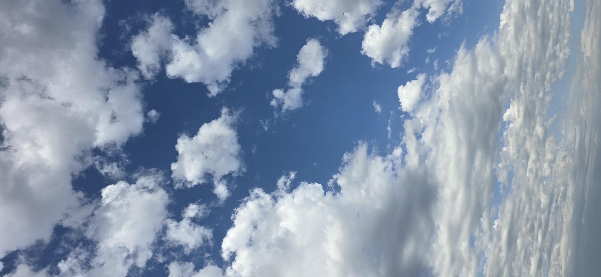 The image depicts a clear blue sky filled with a variety of clouds. These range from small, puffy cumulus clouds scattered across the upper half of the frame to larger, more textured clouds, some appearing wispy and others dense and billowy. Toward the bottom of the image, the clouds become more layered and spread out, with some darker, more ominous-looking formations mixed with lighter, more diffused ones. The overall impression is a dynamic sky with good visibility of the cloud formations against the bright blue backdrop. The scene is devoid of any man-made structures or living beings, suggesting an outdoor environment focused solely on the atmospheric conditions.