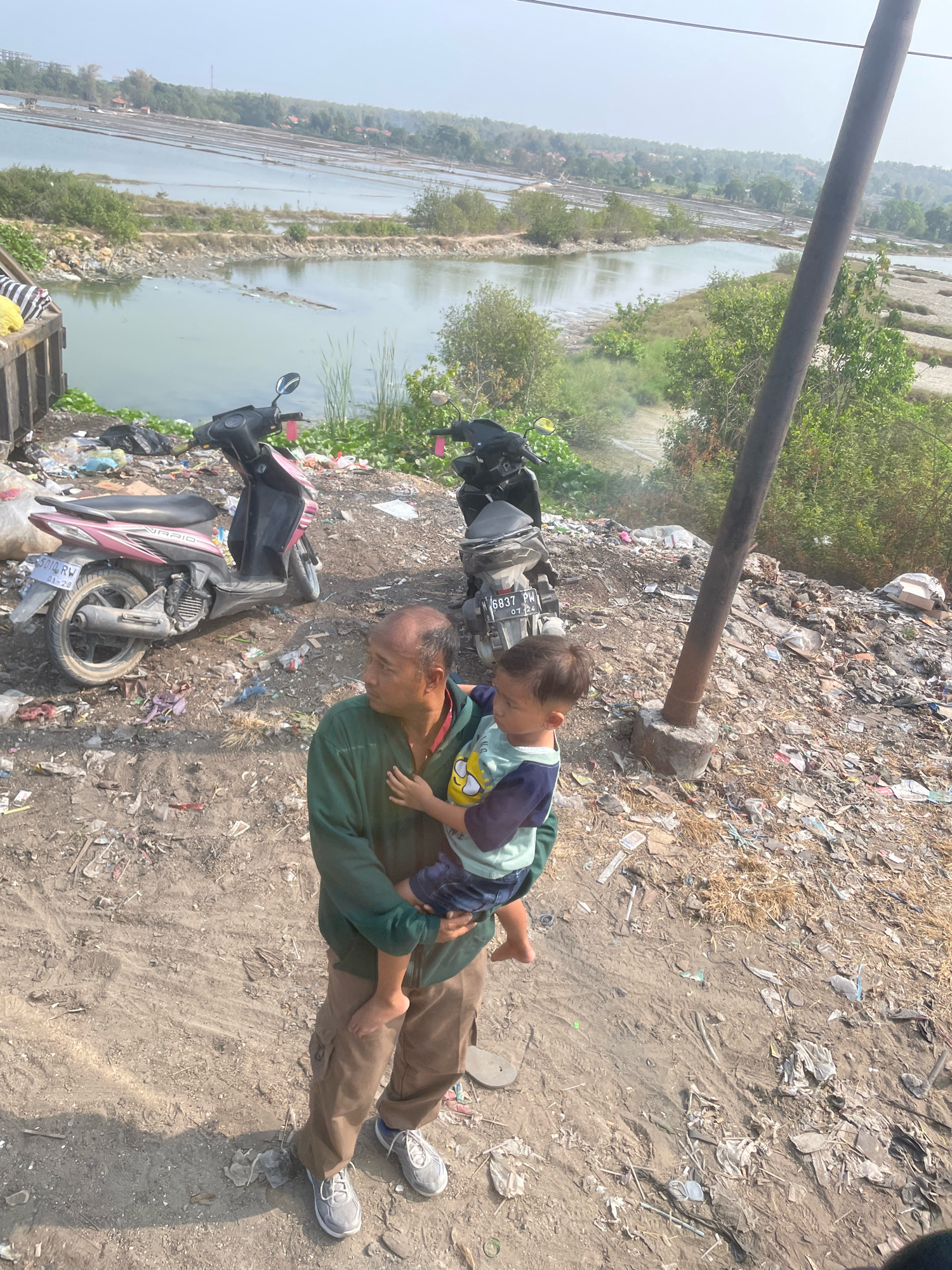 An Indonesian man, likely in his 40s with thinning hair and a weathered complexion, stands on a dirt path. He is wearing a dark green long-sleeved shirt and brown cargo pants. He is holding a young child, a boy around 2-3 years old, who is wearing a light blue and yellow t-shirt and dark shorts. The man is looking away from the camera, towards the right side of the frame. The child is facing the camera.
The background suggests a rural or semi-industrial area. In the distance, there are salt pans or ponds, characteristic of coastal agricultural areas. The immediate foreground is a cluttered area with a significant amount of garbage, including plastic bags, bottles, and other debris scattered across the ground. Two motorcycles are parked near the garbage pile, one pink and white, the other black. A dark wooden pole, possibly for utility lines, stands upright near the center of the frame. The overall environment appears dry and dusty, with sparse vegetation. The sky is hazy, indicating a possibly warm or humid day. The scene is located near the city of Camplong, Indonesia, known for its coastal areas and salt production.