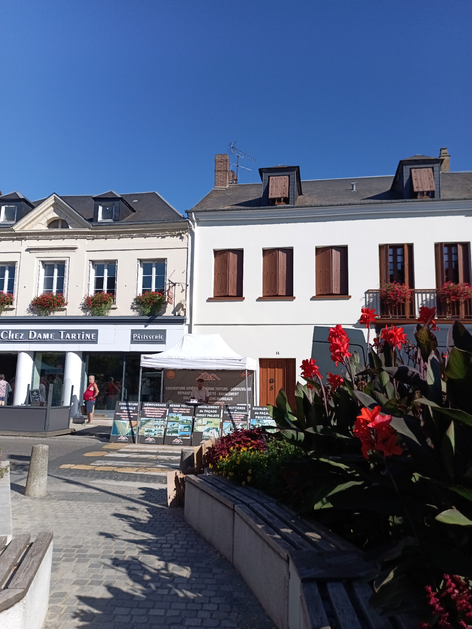 In Le Neubourg, France, on a sunny day, a street scene unfolds. The primary focus is on a building facade featuring two adjacent establishments: "Chez Dame Tartine" and "Pâtisserie". These businesses occupy the ground floor of a light-colored building with traditional French architectural elements, including white-framed windows adorned with red flower boxes. Above the shops, a darker roofline with dormer windows is visible. To the right, a separate white building stands, also with brown-shuttered windows.
In front of the buildings, a temporary white tent is set up, displaying various advertising posters. A man in a red t-shirt and blue shorts stands near the tent, facing the street. Several other people are visible in the background, some entering or exiting "Chez Dame Tartine".
The foreground features a cobblestone street with a pedestrian crossing marked by yellow and white stripes. A concrete bollard stands on the left side of the street. A wooden bench is positioned in the lower right corner, partially obscured by lush, dark green foliage with bright red flowers in bloom. Shadows of trees are cast across the cobblestones, indicating the time of day. The sky is clear and blue.