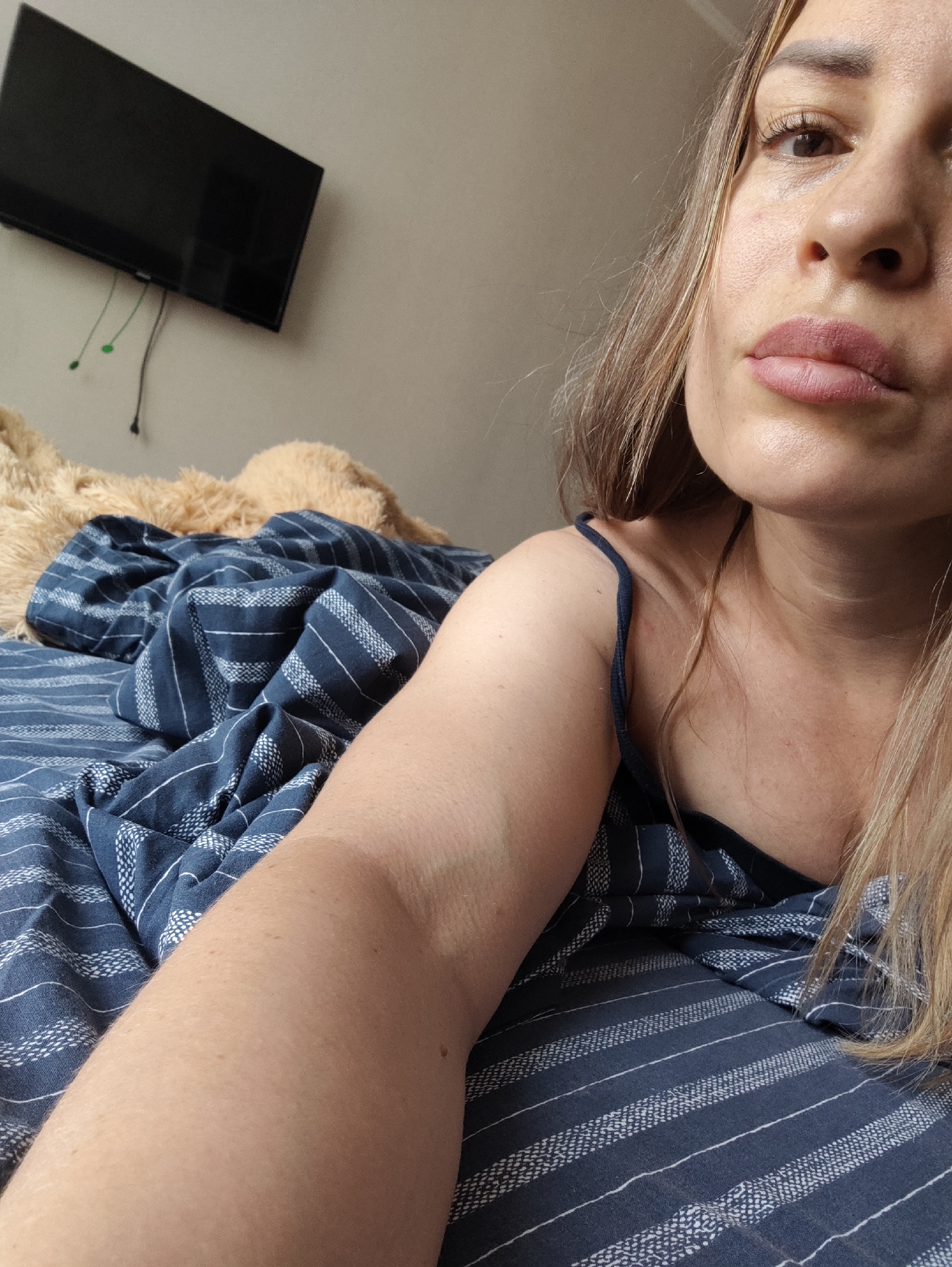 A woman with blonde hair and light skin is lying in bed, partially covered by a dark blue and white striped blanket. She is wearing a dark blue camisole with thin straps. Her face is close to the camera, with her lips slightly parted. Her right arm is visible, resting on the blanket. A large, fluffy, light brown stuffed animal is partially visible behind her. In the background, a black television is mounted on a light gray wall. The scene appears to be an indoor setting, likely a bedroom. There is no indication of the specific location within Almaty, Kazakhstan.