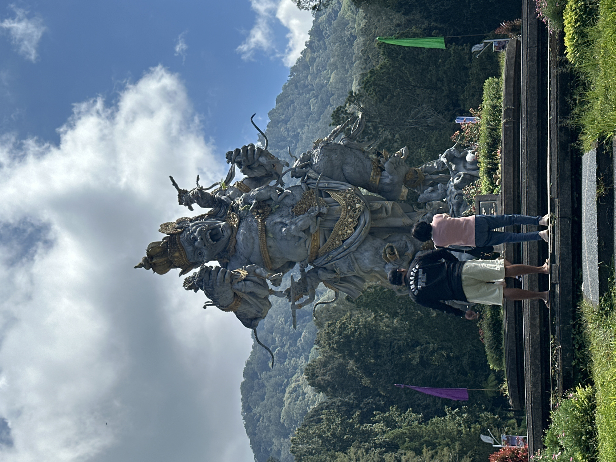 In Baturiti, Indonesia, a large stone statue of a multi-armed mythical figure stands prominently against a backdrop of lush green mountains and a cloudy sky. Two individuals, a man and a woman, are standing on a tiered stone platform facing the statue, their backs to the camera. The man is wearing a black long-sleeved shirt with white lettering and light-colored shorts, while the woman is wearing a pink top and blue jeans. Two flags, one purple and one green, are visible on either side of the platform. The foreground is filled with grass and flowering plants.