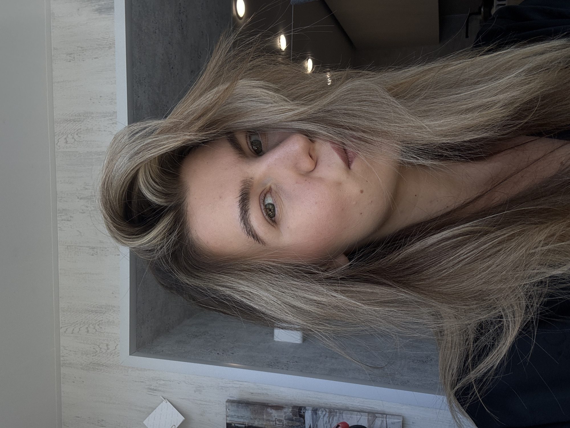 A woman with long, light brown hair is facing the camera. She has light skin, brown eyes, and dark eyebrows. The background consists of a wall with a gray section illuminated by recessed lights above. Below the gray section is a lighter section, and what appears to be artwork is partially visible at the bottom.