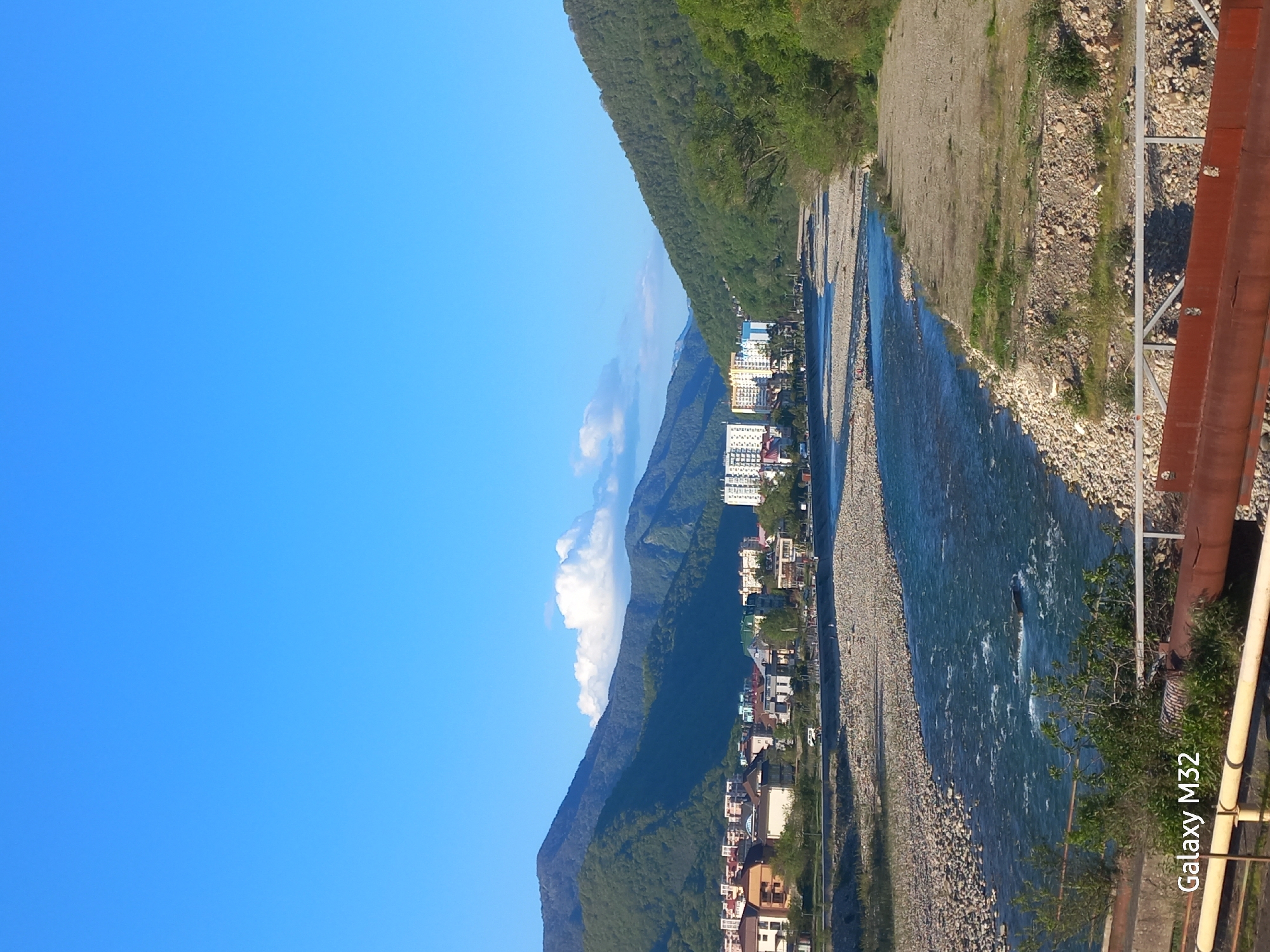 The image shows a mountain river flowing through a town in Sochi, Russia. The river is bordered by rocky banks and vegetation. Buildings are visible along the riverbank. The sky is blue with some clouds. The image appears to be taken from a bridge or elevated position, as evidenced by structural elements of a bridge/railing on the right side of the frame. The camera's name "Galaxy M32" is in the lower right corner.