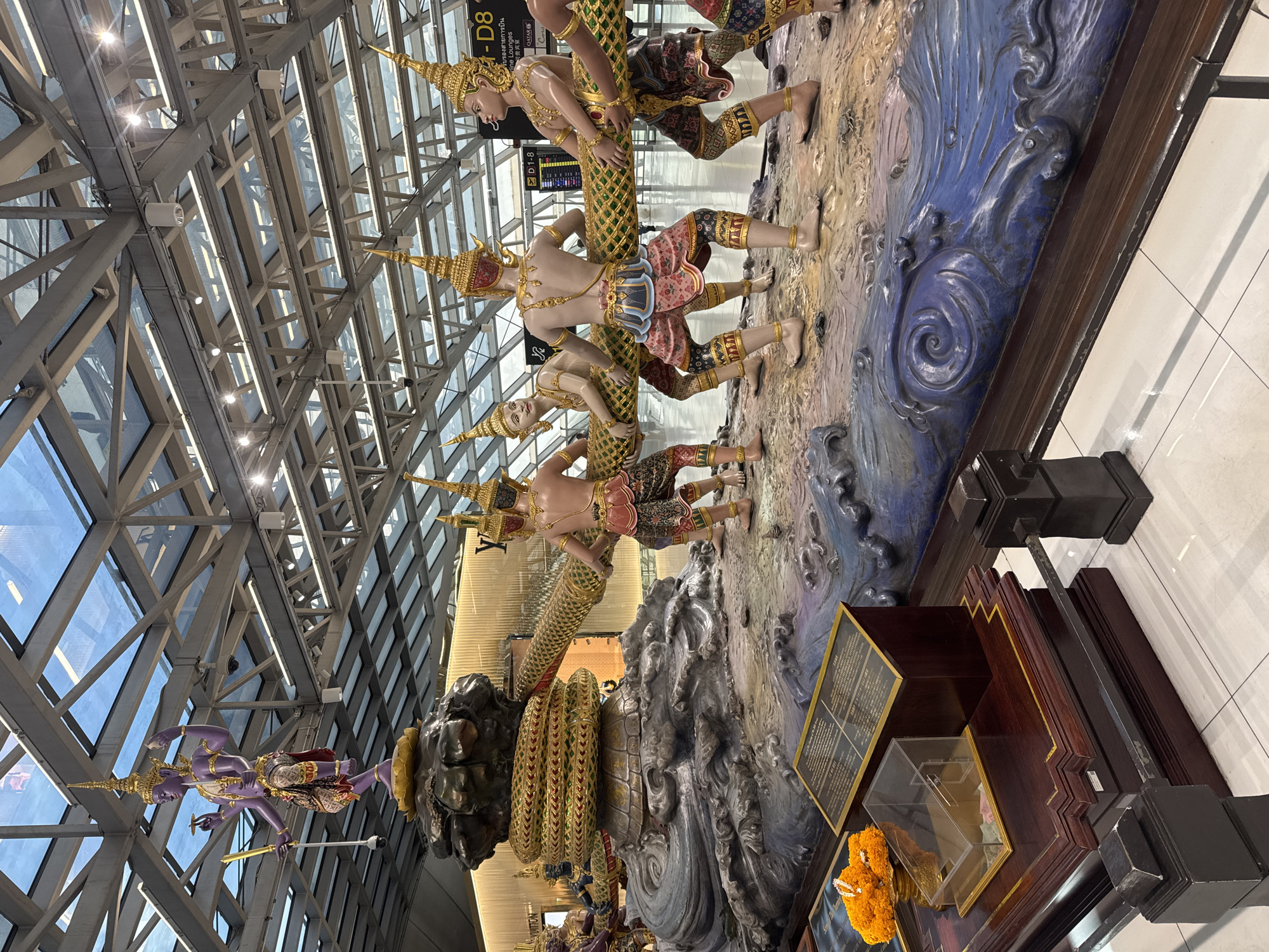 This is a vertical, full-shot photograph of a large, elaborate sculpture depicting figures from Thai mythology, likely located in a public indoor space, such as an airport terminal in Nong Prue Subdistrict, Thailand. The sculpture is vibrantly colored and intricately detailed, featuring several human-like figures wearing traditional Thai headdresses and ornate clothing. These figures appear to be engaged in some form of struggle or ceremonial act, as they are posed dynamically and connected by a large, serpentine creature.
The sculpture is set against a backdrop of what appears to be a stylized representation of water and land, with various textures and shades of blue, brown, and earth tones. The figures themselves are sculpted with realistic human anatomy and adorned with gold jewelry, adding to the richness of the artwork.
Above and behind the sculpture, the architecture of the interior space is visible, characterized by a large, modern glass and steel ceiling structure, indicative of a contemporary building. The lighting suggests it is daytime, with natural light filtering through the expansive windows, supplemented by interior lighting.
Visible text in the image includes what appears to be airport gate information ("D1-8", "- D8"), airline logos ("QATAR"), and possibly signage in Thai and Chinese, reinforcing the context of an international transportation hub. The overall impression is one of cultural artistry and grandeur, presented in a busy, well-lit public environment. The presence of an offering of orange marigold flowers in the foreground, placed near a display case, suggests a cultural or religious significance to the sculpture.