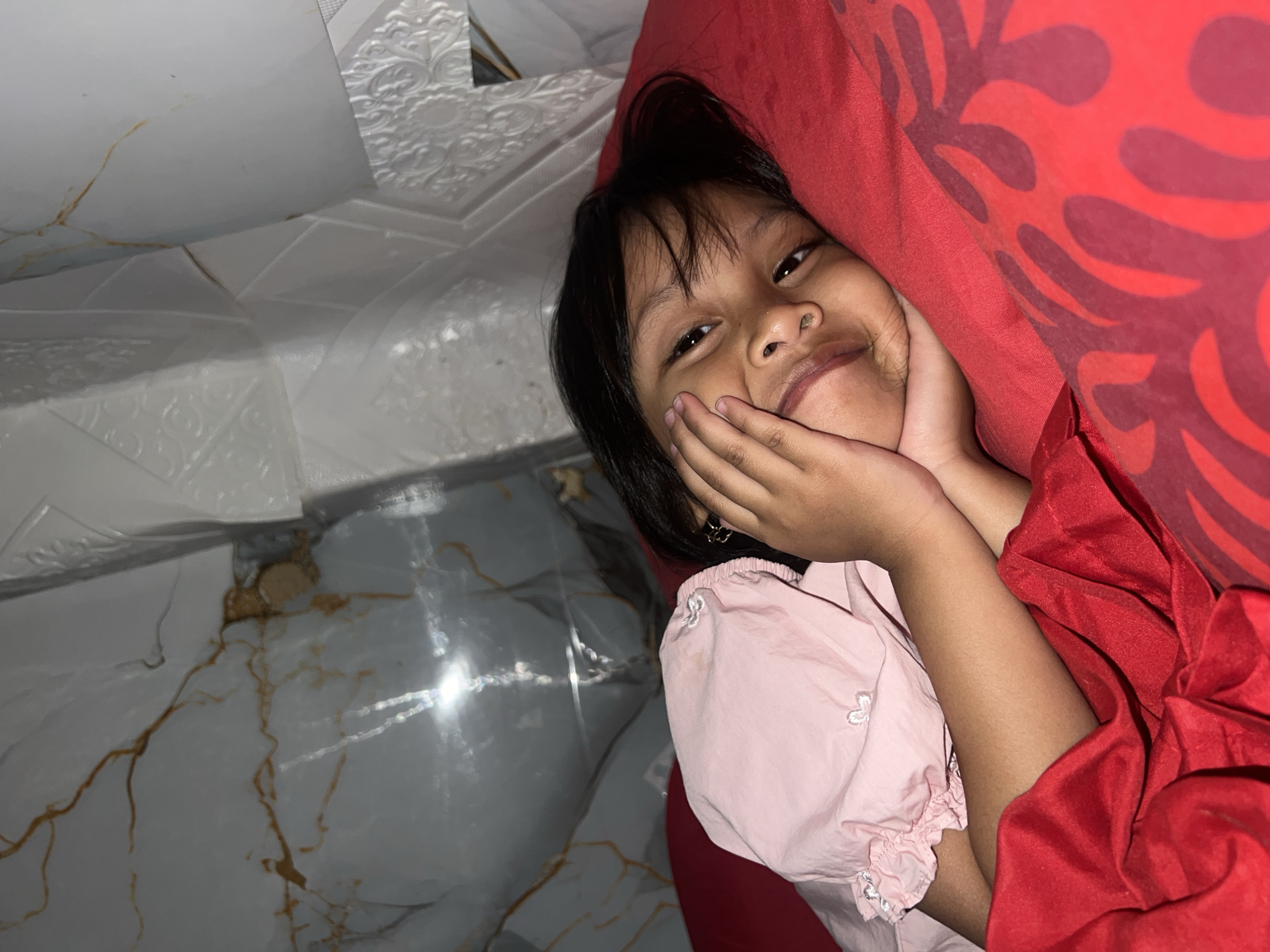 A young girl with dark hair is lying in bed, propped up against a red pillow. Her hands are on her cheeks, and she is smiling at the camera. She is wearing a pink shirt with white embroidery. The room appears to be a bedroom, with patterned wallpaper and a marble-patterned floor visible. The lighting suggests it might be nighttime or indoors with artificial light. The girl's expression is happy and playful. No text is visible in the image. The image was taken in Kecamatan Pati, Indonesia.