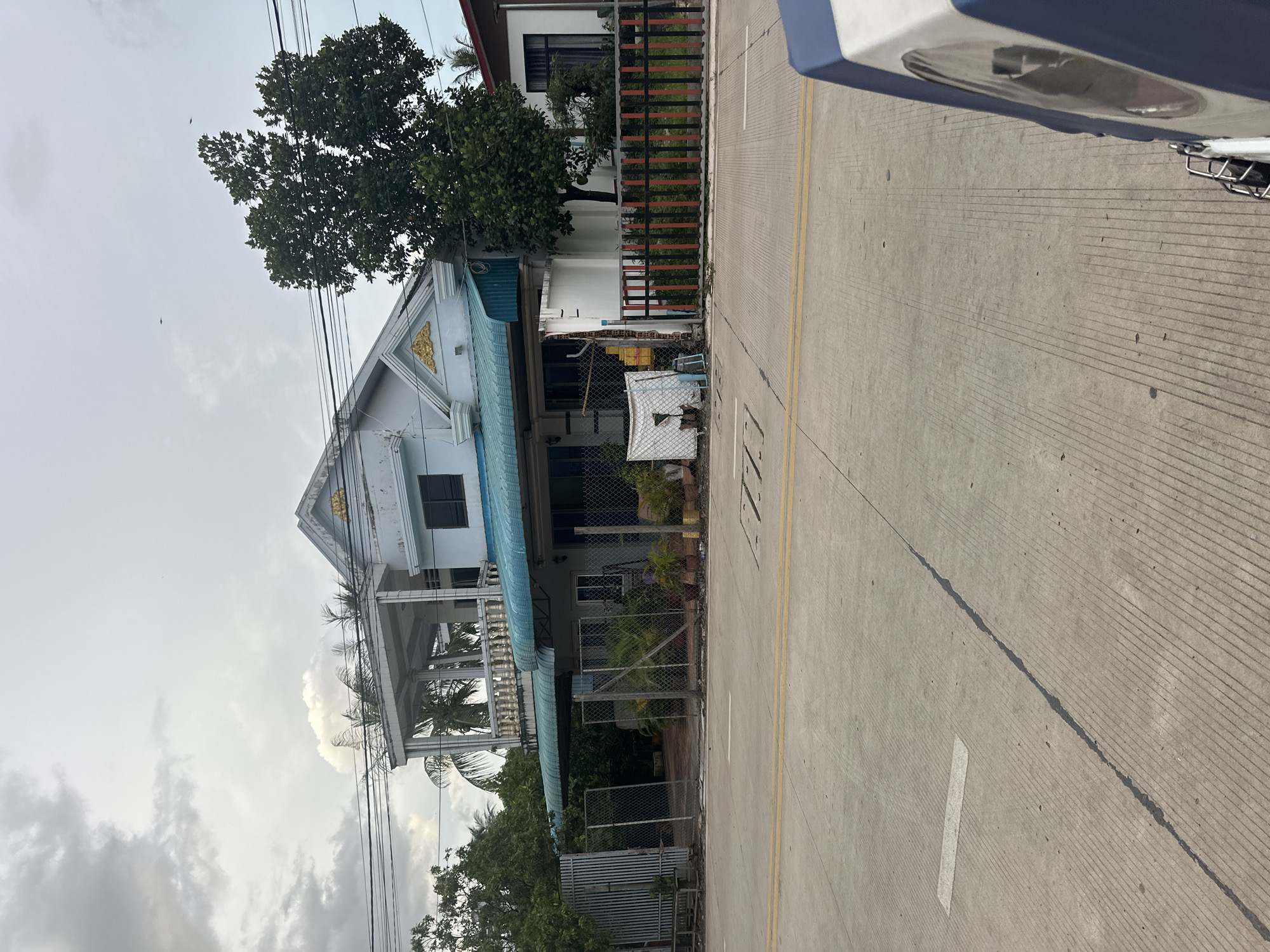The image shows a light blue, two-story building with white trim, likely a residence, located on a street in Kampot, Cambodia. The building has a prominent balcony on the second floor with ornate railings and columns. A teal-colored awning extends over the ground floor, and the facade is decorated with gold accents. The property is partially enclosed by a chain-link fence and a decorative fence with vertical red and black slats. Several potted plants are visible in the foreground and within the fenced area. The street in front of the building is paved with concrete and has yellow lane markings. The sky is overcast with gray clouds. There are no people visible in the scene, and the overall impression is that of a quiet residential street.