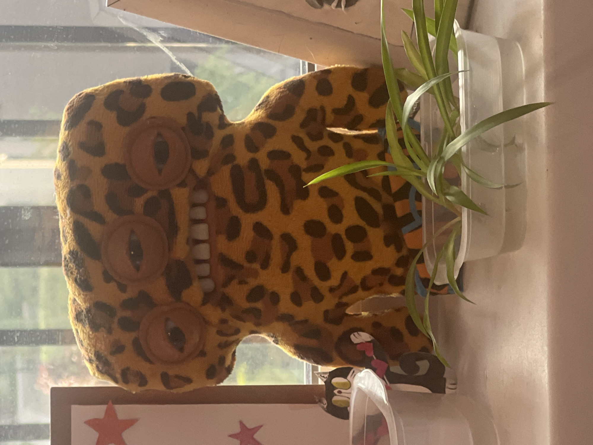 The image displays a still life arrangement taken indoors, oriented vertically but depicting subjects that would naturally be viewed horizontally. In the center, a plush toy with a distinct leopard print pattern is visible. This toy has a creature-like appearance, featuring three large, oval-shaped brown eyes with dark pupils, and a prominent row of white, human-like teeth across its "mouth" area. The plush is made of a soft, textured material, and its body is somewhat shapeless, suggesting a decorative or cuddly item.
To the right of the leopard plush, a plant with long, slender green leaves, possibly a spider plant, is housed in a clear, rectangular plastic container. The container is translucent, allowing some light to pass through. Behind the plant and plush, a window is visible, showing a blurry outdoor background with hints of green foliage, indicating daylight.
Below the plush toy, on what appears to be the surface it's resting on, a small black and white cat figurine or cutout with prominent yellow eyes is partially visible. Further to the bottom left, a section of a framed item can be seen, containing a red star against a white background.
The lighting in the scene is natural, coming from the window, suggesting it is daytime. The overall impression is a quirky collection of items arranged on a windowsill or shelf. There is no discernible activity taking place, and no text is visible in the image. There are no specific visual cues within the image that confirm the location as Saint Petersburg, Russia.