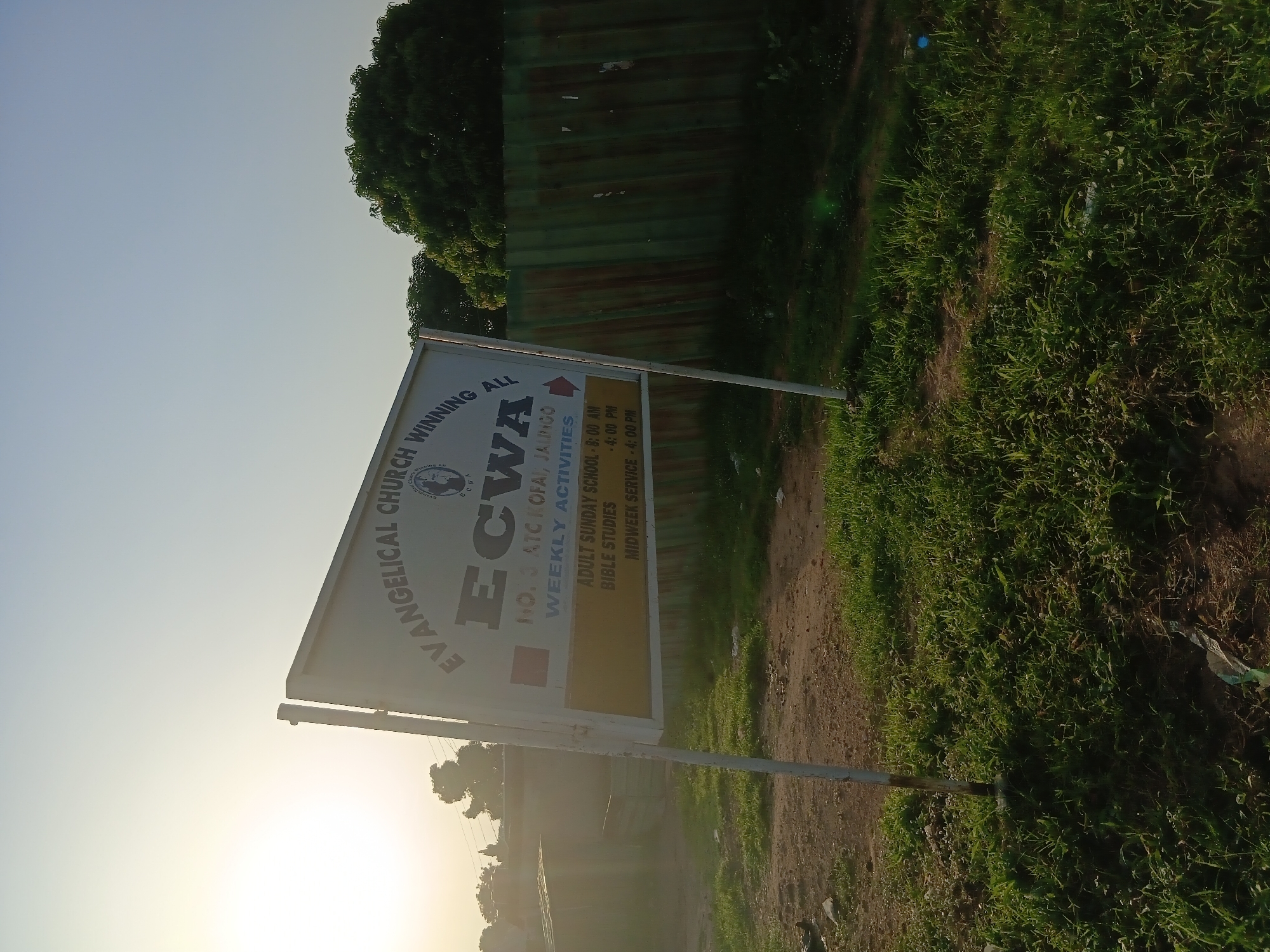 The image shows a sign for the Evangelical Church Winning All (ECWA) located in Jalingo, Nigeria. The sign, mounted on a white metal pole, is white with black lettering. The top of the sign reads "EVANGELICAL CHURCH WINNING ALL" with a circular logo featuring a globe and the text "ECWA" above and below. Below this, in larger font, is "ECWA". Further down, it states "NO. 3, ATC KOFAI, JALINGO". A red arrow points to the right. Below this, on a yellow background, it lists "WEEKLY ACTIVITIES", followed by "ADULT SUNDAY SCHOOL - 8:00 AM", "BIBLE STUDIES - 4:00 PM", and "MIDWEEK SERVICE - 4:00 PM". The sign is positioned in an outdoor environment with green grass and dirt in the foreground. In the background, there are some buildings and a green corrugated metal fence. The sky is a pale blue with the sun shining brightly, casting a warm glow on the scene and creating lens flare. The overall impression is of a sunny day in a semi-urban or rural setting.