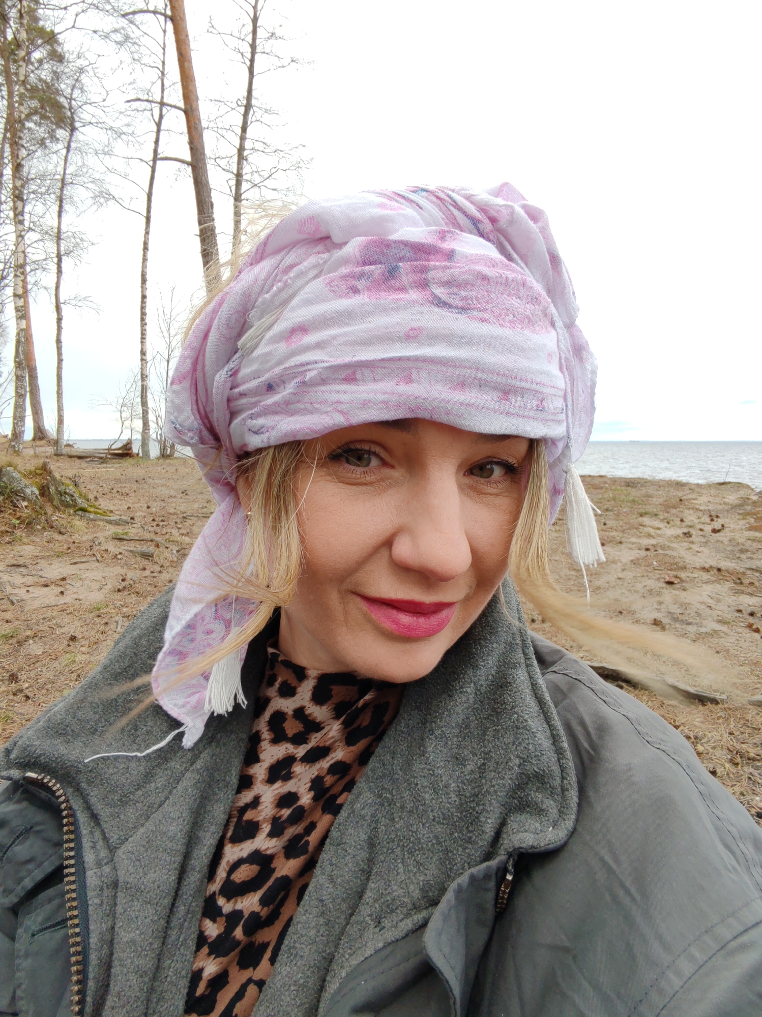 A middle-aged woman is taking a selfie outdoors, likely in Bolshaya Izhora, Russia, given the location name. The background features a sandy beach, bare trees, and a body of water under an overcast sky. The woman is wearing a pink and white patterned headscarf, a leopard-print top, and a grey jacket. She has blonde hair and is wearing pink lipstick. The scene appears natural and unposed.