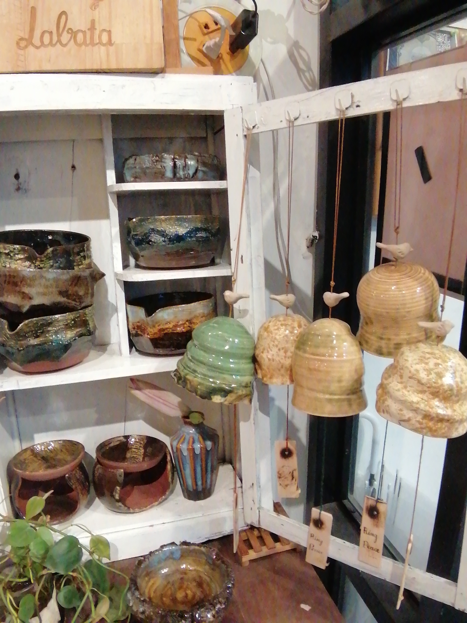 The image shows an indoor display of handcrafted ceramic items, likely in a shop or studio. The items are arranged on white shelves within an open white cabinet. On the top shelf, there are two large, irregularly shaped bowls. Below them, on the second shelf, are three more bowls, two of which are notably wider and shallower than the one above. The third shelf displays a stack of three more wide, shallow bowls, each with a textured, glazed finish in earthy tones of brown, green, and blue.
To the right of the cabinet, an open white wooden frame is visible, from which several ceramic items are hanging by thin cords. These hanging items appear to be bell-shaped or rounded, with small ceramic birds perched on top of some of them. Some of these hanging items have small wooden tags attached with inscribed text, one of which is legible as "Ring Peace".
In the foreground, on a wooden surface, there are two more ceramic bowls, one dark brown and the other a reddish-brown. A leafy green plant is visible to the left, its vines reaching towards the bowls. Further in the foreground, another ceramic item with a textured, iridescent surface is partially visible.
A wooden sign with the word "Labata" in cursive script is mounted above the cabinet. In the background, a window offers a partial view of an outdoor scene or another room, with indistinct light sources and reflections. The overall impression is one of a curated display of artisanal pottery. The location is described as Unknown, Unknown.