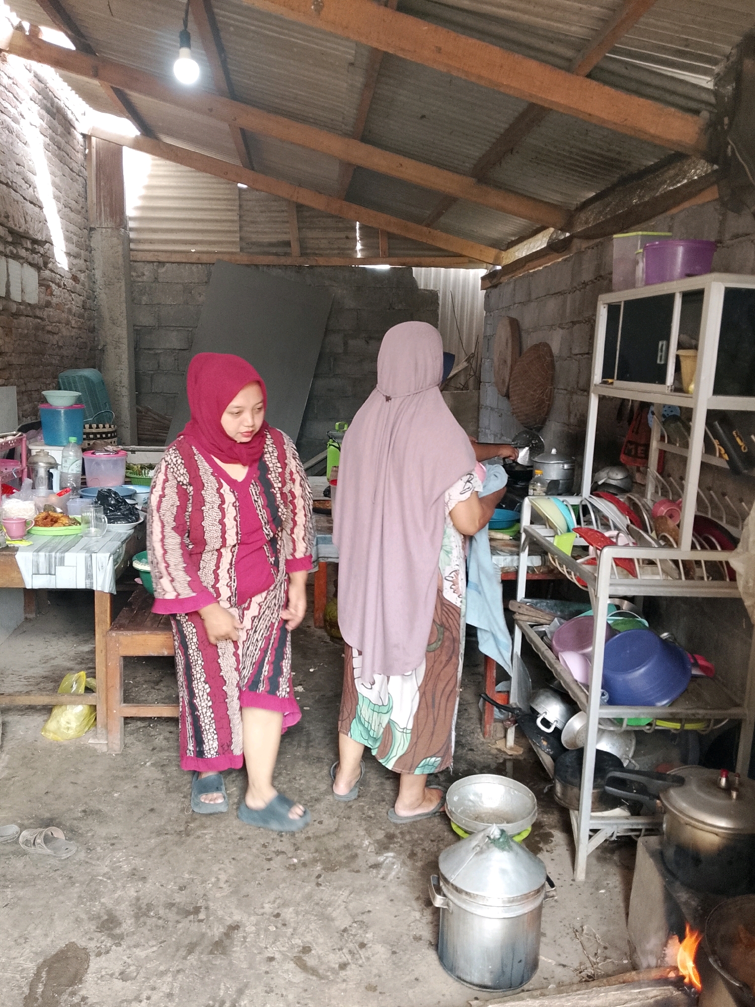 In Karangsari, Indonesia, two women are present in a rustic, open-air kitchen. The woman on the left, wearing a maroon hijab and a patterned dress with shades of maroon and beige, stands facing slightly to the right, looking downwards. The woman on the right, draped in a pale pink hijab and a patterned skirt with green and brown hues, faces away from the camera and is engaged in an activity near a cooking area.
The environment appears to be a humble dwelling with a corrugated metal roof supported by wooden beams. The walls are constructed from cinder blocks and exposed brick. The floor is concrete. Various kitchen items are scattered throughout the scene, including pots, pans, bowls, and containers, some of which are stacked on a metal shelving unit. A stove with a visible flame is situated in the lower right portion of the image, indicating active cooking. The overall impression is one of a functional, lived-in kitchen space.