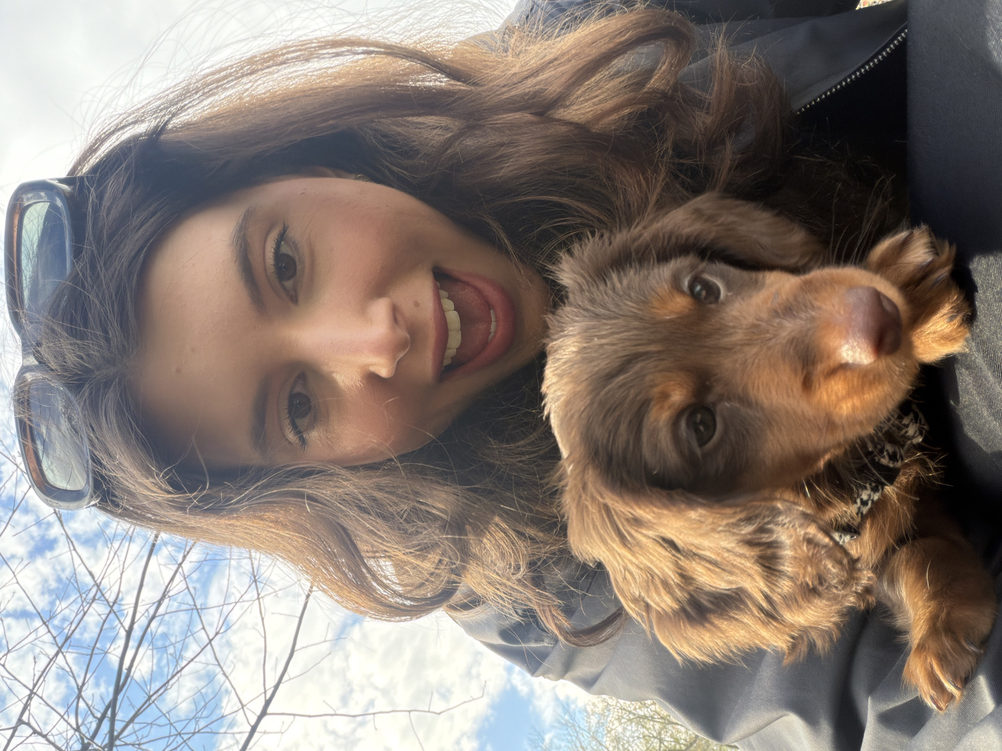 A young woman with long, brown hair and sunglasses on her head poses for a selfie with a brown puppy with floppy ears. The woman is smiling broadly with her tongue slightly visible. The puppy looks at the camera. They are outside on a partly cloudy day with bare tree branches visible in the background. The exact location in Montreal, Canada cannot be determined from the image.