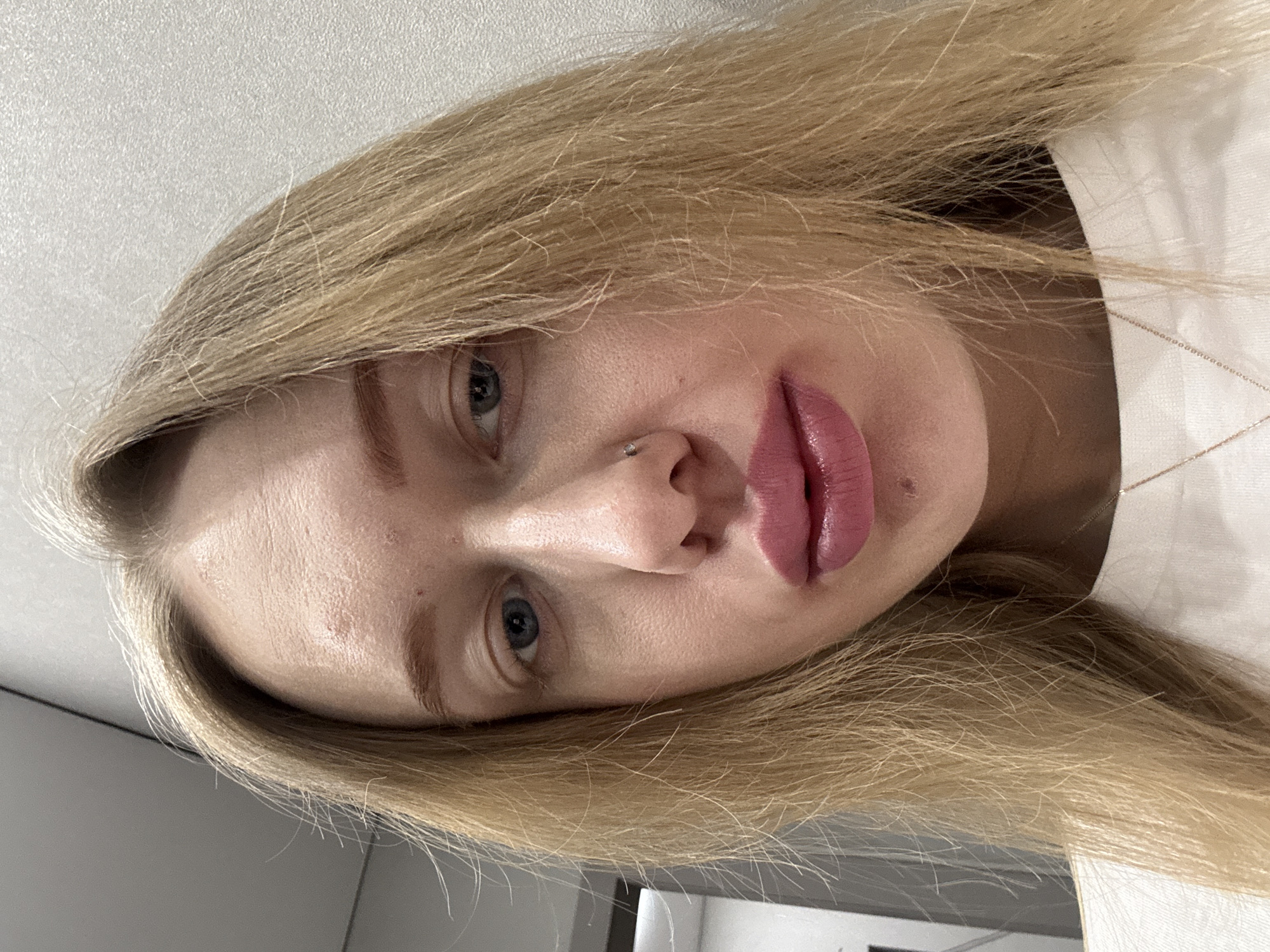 A close-up, slightly angled portrait shows a young woman with fair skin, blonde hair, and light-colored eyes. She has a nose ring and full, pink lips. She is wearing a white garment, with a thin gold necklace visible at her collarbone. The background includes a plain white surface and a glimpse of a gray surface, possibly a wall. There is no information available to determine the location in the city of Novoaleksandrovka, Russia.