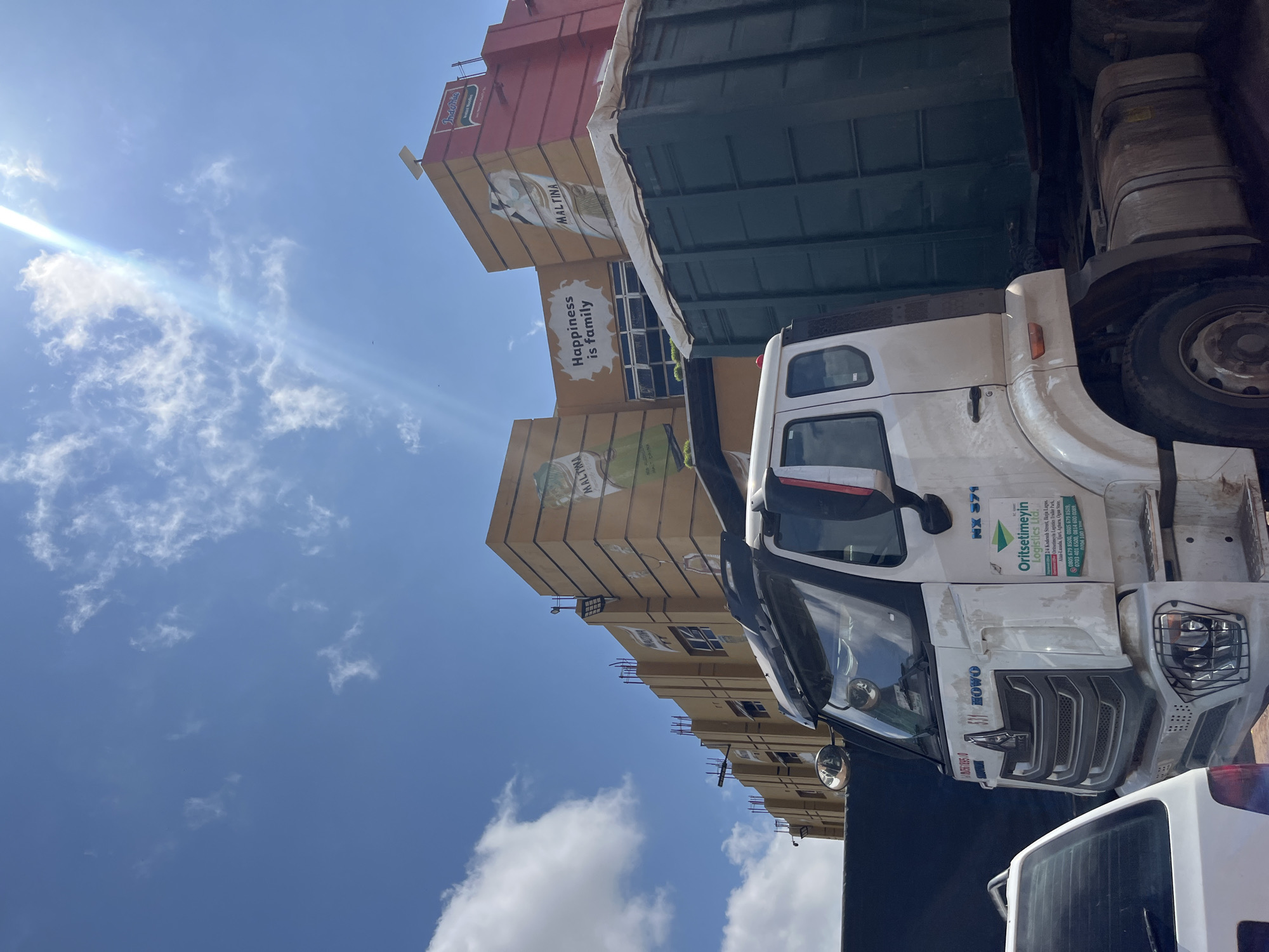 The image depicts a street scene in Jos, Nigeria, dominated by a white semi-truck parked in front of a building. The building, painted in shades of yellow and red, features prominent signage for "MALTINA" and an advertisement that reads "Happiness is family." The sky is blue with scattered white clouds, and the sun is bright, creating lens flare. The truck has "Oritselimeyin Logistics Ltd." written on its side, along with contact information. A portion of a dark green trailer is visible behind the truck, and the rear of a white car is in the foreground on the left. The overall impression is of a commercial area on a sunny day.