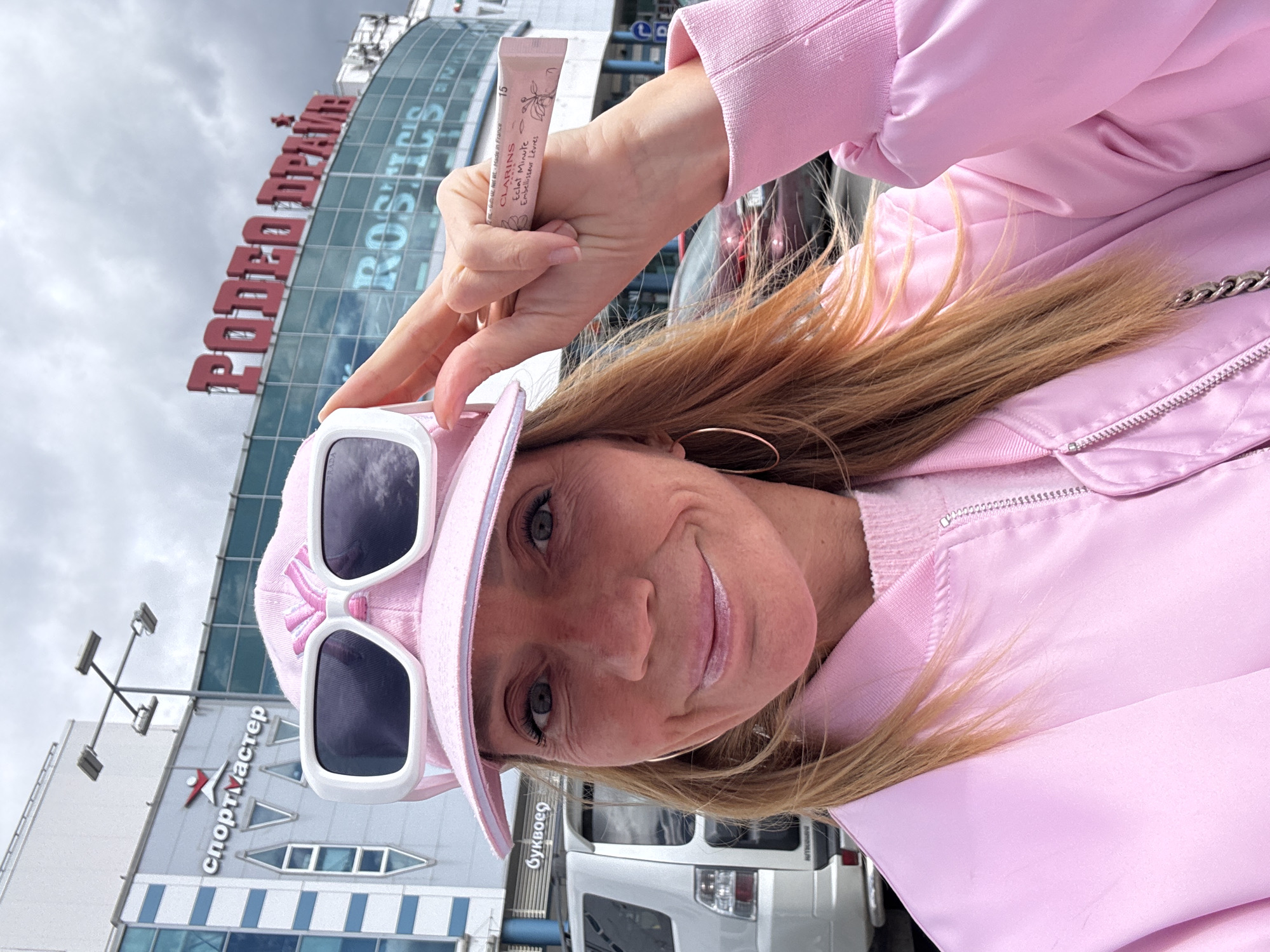 A woman in a pink outfit, including a cap and jacket, holds a tube of Clarins lip gloss in front of her. She wears white-framed sunglasses and hoop earrings. In the background is a building with "спортмастер" written on it, and another building with "РОДЕО ДРАЙВ" above "RODEO DRIVE" in Cyrillic. A white van is visible in the background as well. This location is in Saint Petersburg, Russia.