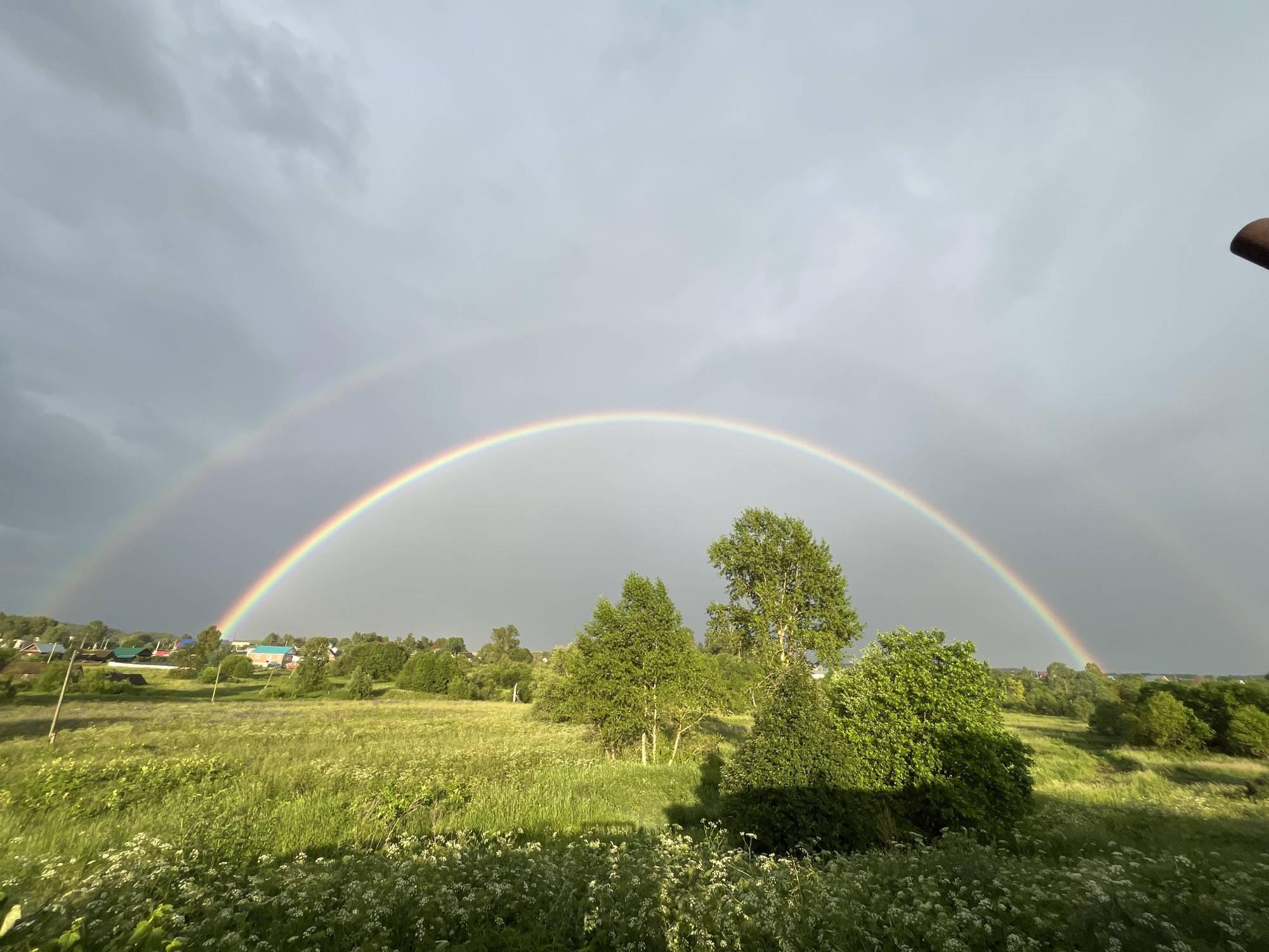 This media file depicts a serene rural landscape in Уральское, Russia, under a dramatic sky. The primary subjects are a vibrant double rainbow arcing across the frame, set against a backdrop of lush green fields and scattered trees. In the distance, small houses with colorful roofs suggest a village or settlement. The foreground is filled with tall grasses and delicate white wildflowers, adding a touch of natural beauty to the scene.
The time of day appears to be late afternoon or early evening, indicated by the soft, diffused light and the storm clouds that are beginning to dissipate, allowing the sun to break through and create the rainbows. The weather is transitioning from rain to clear, with heavy, dark clouds in the upper portions of the sky contrasting with the brighter areas where the rainbows are visible.
Notable details include the clear definition of both the primary and secondary rainbows, the diverse textures of the vegetation, and the distinctly Russian architectural style of the distant houses. The overall mood is one of peace and natural wonder, evoking a sense of calm and awe inspired by the majestic display of the double rainbow. There is no visible text in the media file.