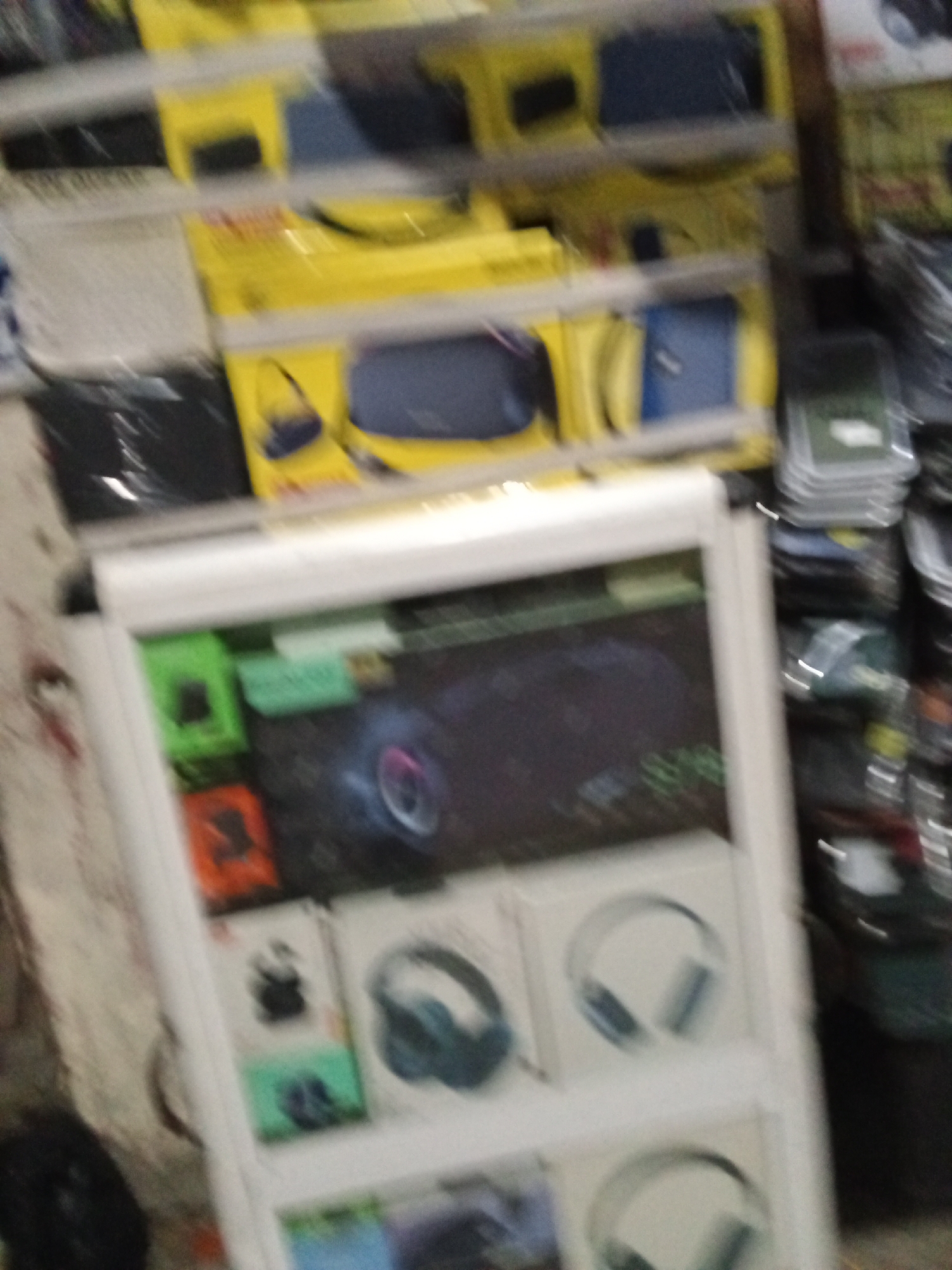The image shows a display of electronic accessories inside a shop, possibly in Ado, Nigeria, judging from the likely setting. The display consists of stacked shelves of packaged items on the top shelf and a white display stand below. The display stand shows various products, including computer mice and headphones, as shown in the images on the product boxes. The shop seems to be stocked with phone chargers, cables, and other electronics. The image quality is blurry, making detailed identification difficult.