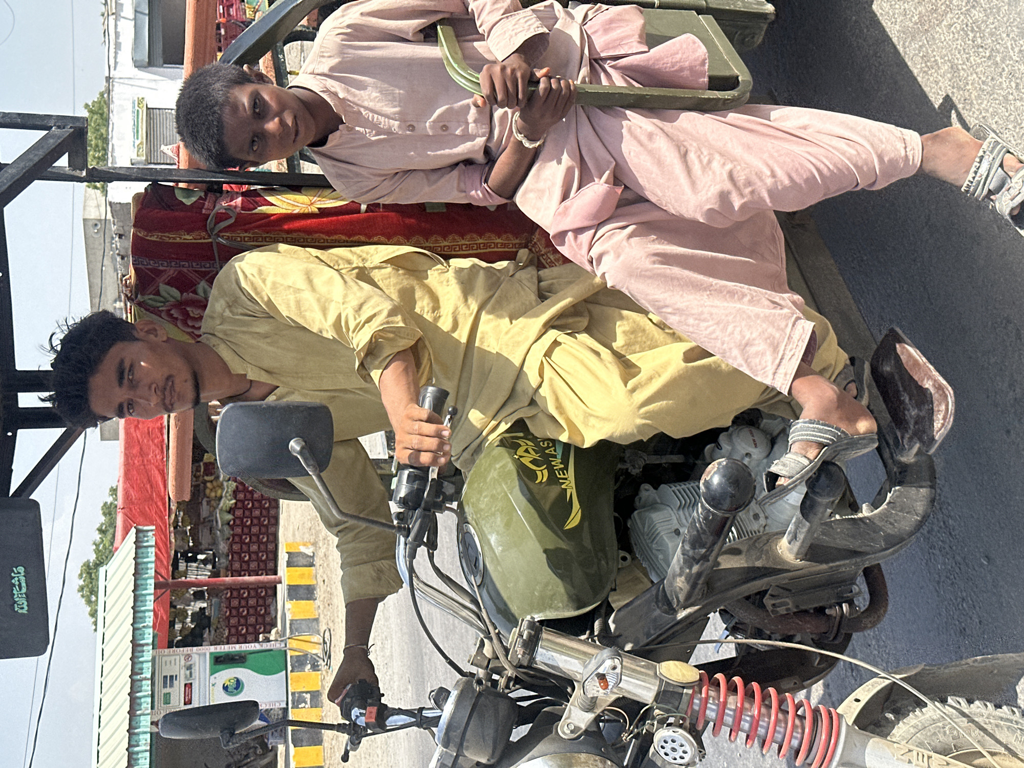 Two males are captured in a daytime, outdoor setting, riding what appears to be a three-wheeled motorcycle taxi or a motorcycle with a sidecar attachment. The image is taken from a slightly elevated perspective, looking down at the subjects.
The individual in the foreground, acting as the driver, is an adult male with light brown skin and dark hair. He is wearing a light yellow traditional garment, possibly a shalwar kameez. His hands are on the motorcycle handlebars, and he is looking directly at the camera with a neutral expression.
Behind the driver, situated in the sidecar or passenger section, is a younger male. He also has light brown skin and dark, curly hair. He wears a light pink traditional outfit, similar to the driver's, and has a silver-colored bracelet on his right wrist. He is holding onto a dark metal bar and is also looking towards the camera. The seating area in the sidecar is covered with a decorative red and yellow patterned fabric or carpet.
The motorcycle itself is olive green, with "NEW AS" and some graphic elements visible on its fuel tank. A black panel on the motorcycle bears the Arabic inscription "ما شاء الله" (Masha'Allah). The front suspension has prominent red springs.
The background suggests a street or urban environment. Buildings are visible, and to the left, there's a structure with a green and white striped awning. Further back, yellow and black striped barriers are on the ground, and a green dispenser, possibly a fuel pump, is visible. Text on the pump includes "YOUR METER 0000 BEFORE D". Piles of what appear to be fruits are seen under a red canopy in the distance, indicating a market stall or shop. The bright lighting suggests it is daytime.