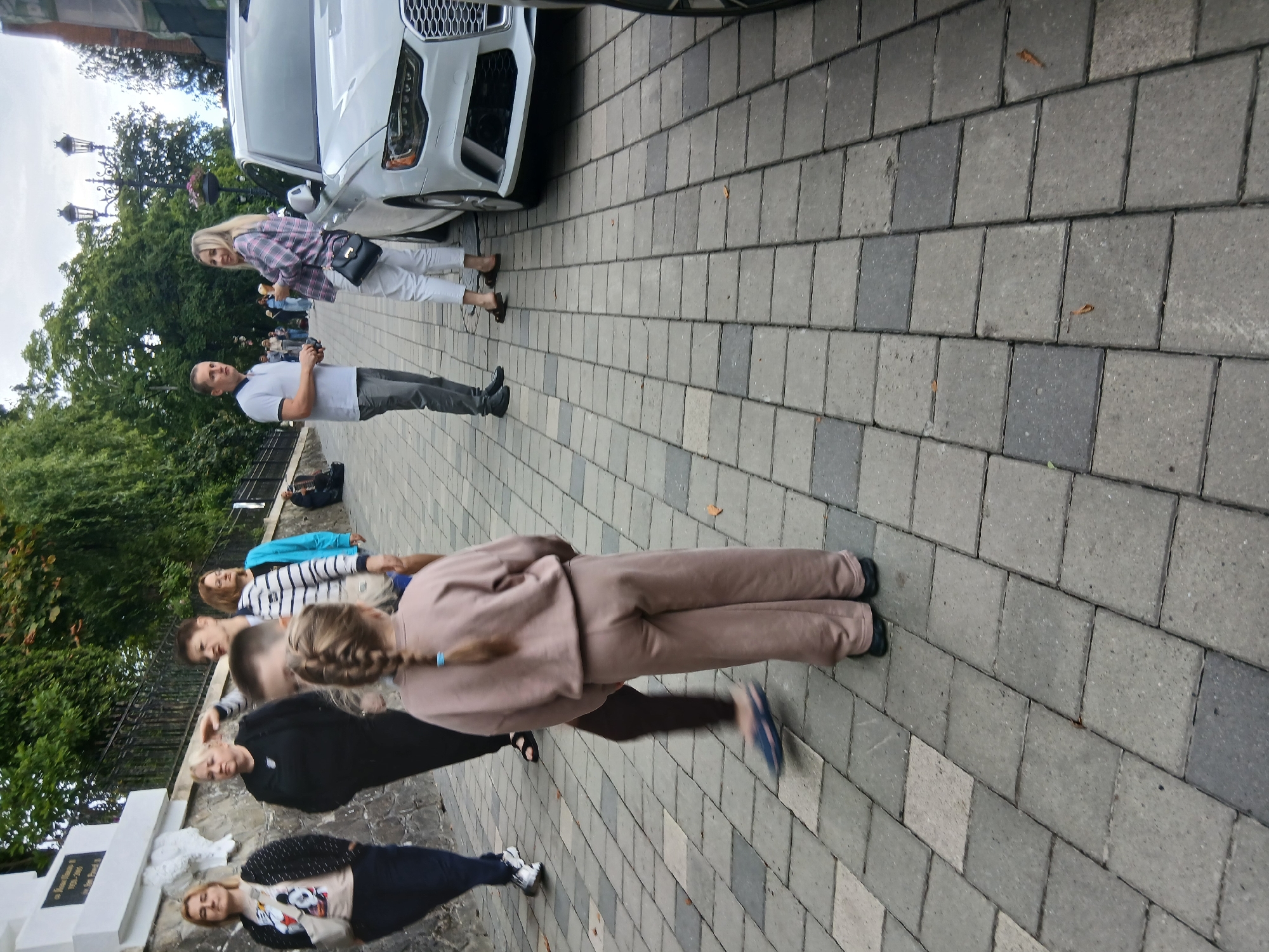 The image captures an outdoor scene on a paved walkway in Truskavets, Ukraine, featuring a group of individuals and a white car. In the foreground, a young girl with blonde hair in a braid is seen from the back, wearing a light brown tracksuit. To her left, another person's legs are visible, wearing blue sandals, suggesting movement.
Further back, several adults are present. On the left, a woman in a black top and dark pants stands with her back partially turned. Next to her, another woman wears a black top. A third woman, wearing a striped top and a blue jacket, is also visible. In the middle ground, a man in a white polo shirt and grey pants stands and holds a phone. Beside him, a woman with blonde hair wears a pink and grey plaid shirt and white pants, carrying a black handbag.
A white car, possibly an Audi, is parked on the right side of the image. The background shows a stone wall on the left, with trees and greenery beyond it. Street lamps are visible in the distance. The walkway is constructed of interlocking grey and darker grey paving stones. The overall atmosphere suggests a casual public gathering or a stroll in a park or street in Truskavets.