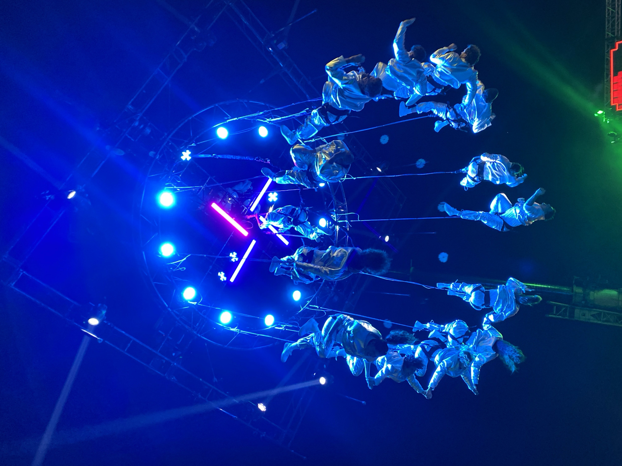 The image shows a group of acrobats suspended in the air on cables, performing in what appears to be an indoor arena. The lighting is dramatic, with deep blues, some greens, and pinks. The acrobats wear light-colored clothing. Above and around them are lighting fixtures, including spotlights. The overall setting is an event or performance, possibly a concert or circus. I don't see any indications of the location being in Buenos Aires, Argentina.