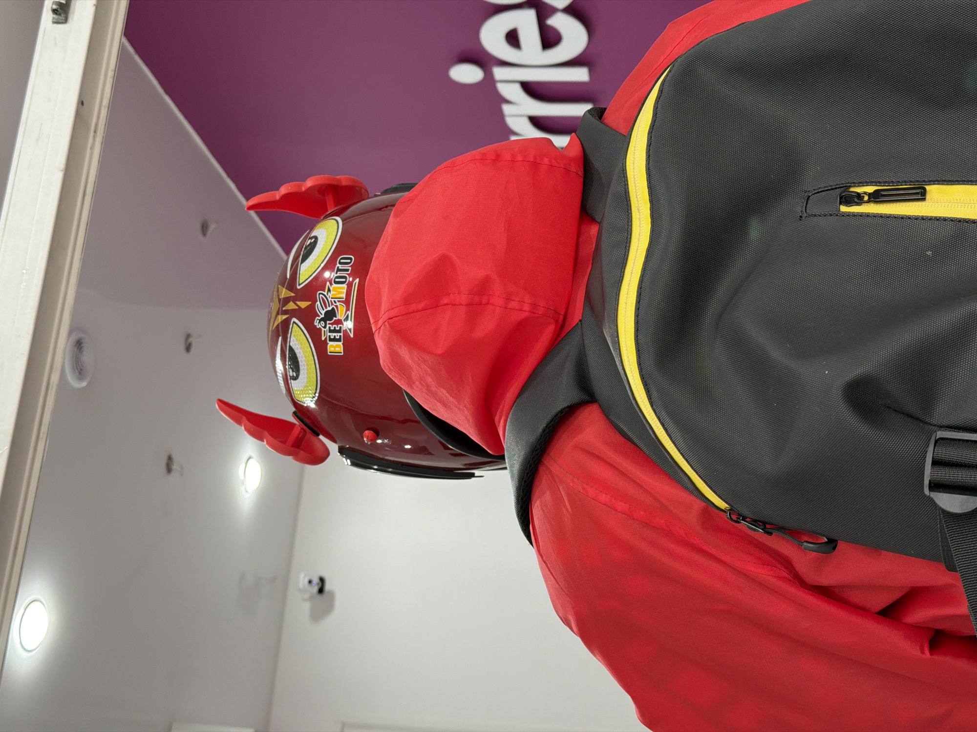 A low-angle, close-up shot focuses on a person in a red hooded jacket and a black backpack with a yellow stripe, and a red helmet with devil horns. The helmet features cartoonish eyes and a logo that reads "BEE MOTO". The person is standing indoors, near a white wall and a purple ceiling with white letters that read "Berries". The setting suggests a business establishment, potentially in Saint Petersburg, Russia, given the Cyrillic alphabet and architectural elements that may be common in that region.