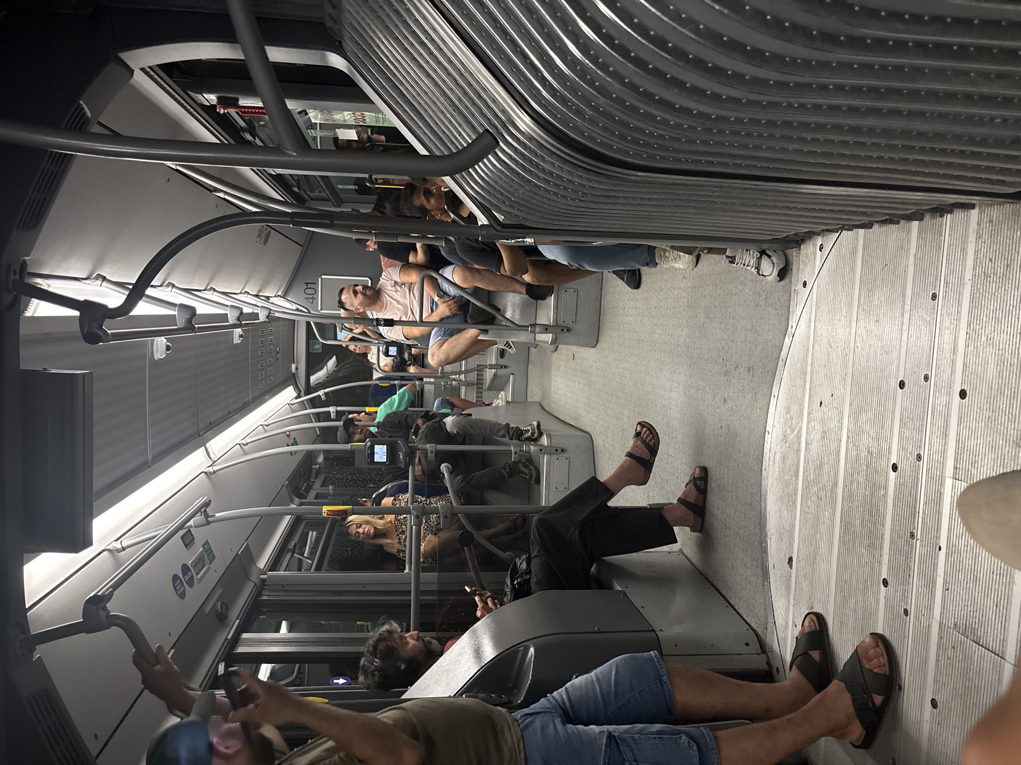 An interior view of a public bus shows several passengers seated and standing. The bus appears to be in motion, suggested by the blurred scenery visible through the windows and the slight motion blur on some of the passengers. The lighting is dim, with overhead fluorescent lights illuminating the aisle and seats. Passengers are dressed in casual summer clothing. One man in the foreground is wearing denim shorts and a green t-shirt and is looking down at his phone. To his left, a man with a beard wearing black pants and sandals is also looking at his phone. Further down the aisle, a woman in a leopard print top is seated. At the front of the bus, a sign displays the number "401". The seating arrangement consists of individual seats and bench-style seating. The floor is made of ribbed metal. The bus has handrails and poles for passengers to hold onto. The scene suggests a typical commute or journey on public transportation. The context of Venice, Italy is not evident from the image itself.