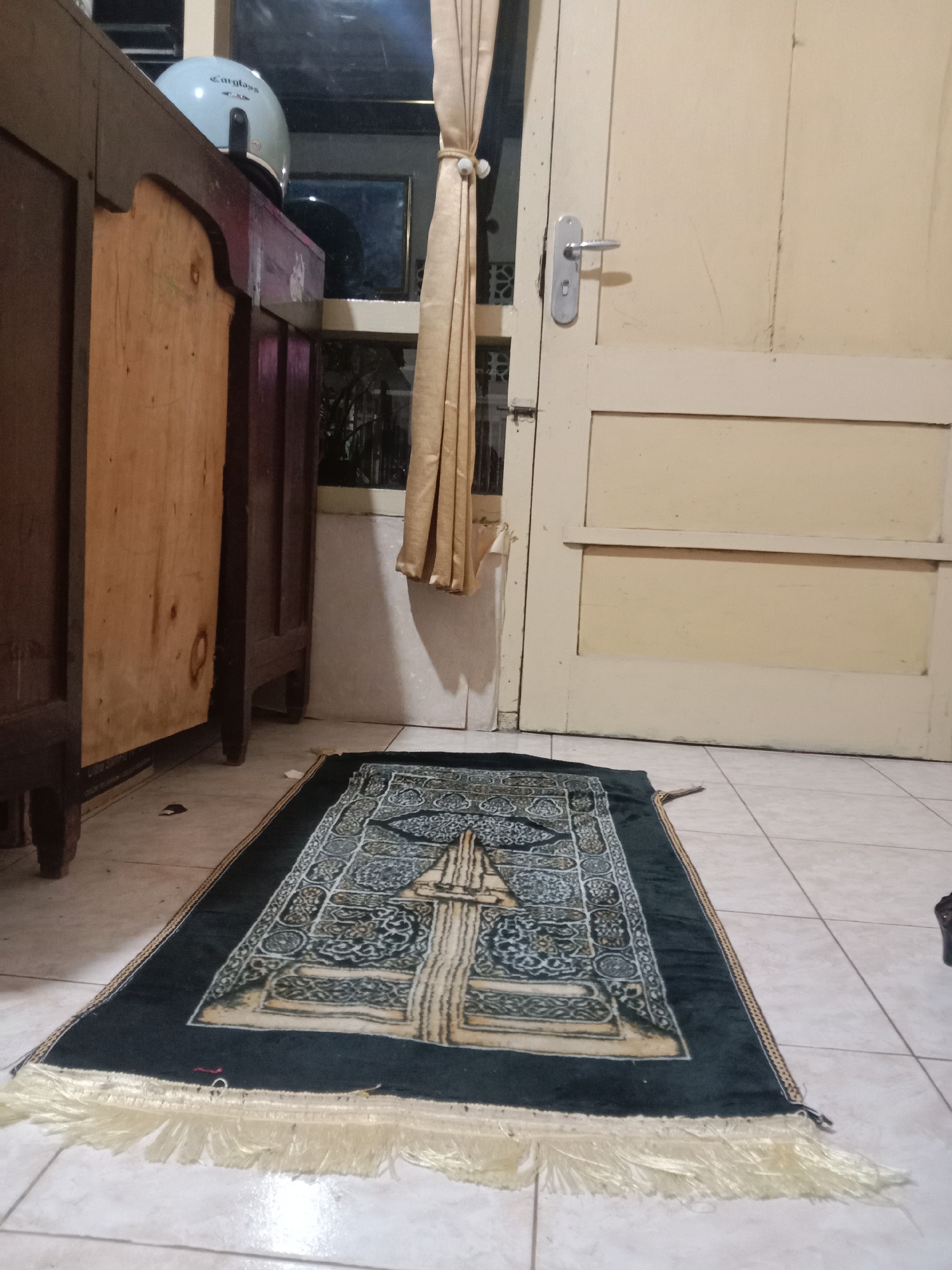 A light blue motorcycle helmet with "Cargloss" written on it rests on top of a dark brown wooden cabinet. To the right of the cabinet is a beige wooden door with a silver handle. In front of the door, on a tiled floor, is a dark green prayer rug with intricate gold and white patterns and a fringe. The lighting in the room is dim, suggesting it may be evening or night. The scene is indoors, in a residential setting, possibly in Madiun, Indonesia, given the cultural context of the prayer rug.