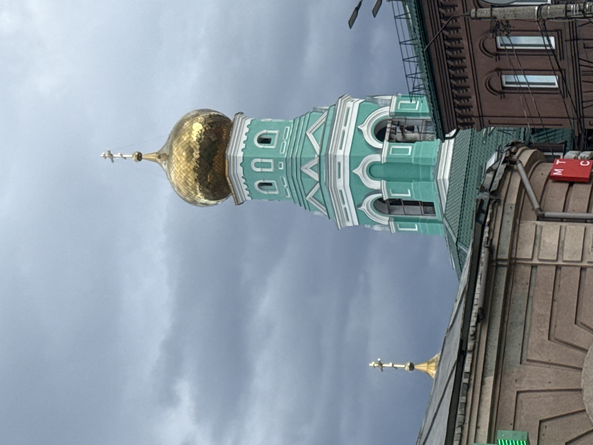 The image shows the tower of the Kazan Cathedral in Syzran, Russia, against a cloudy sky. The tower is light green with white architectural trim and is topped by a gold dome and a cross. Adjacent to the tower are other buildings, including one with brown brick and visible windows, as well as a building with a gray stone facade and a red sign with white Cyrillic letters "MTC" on it.