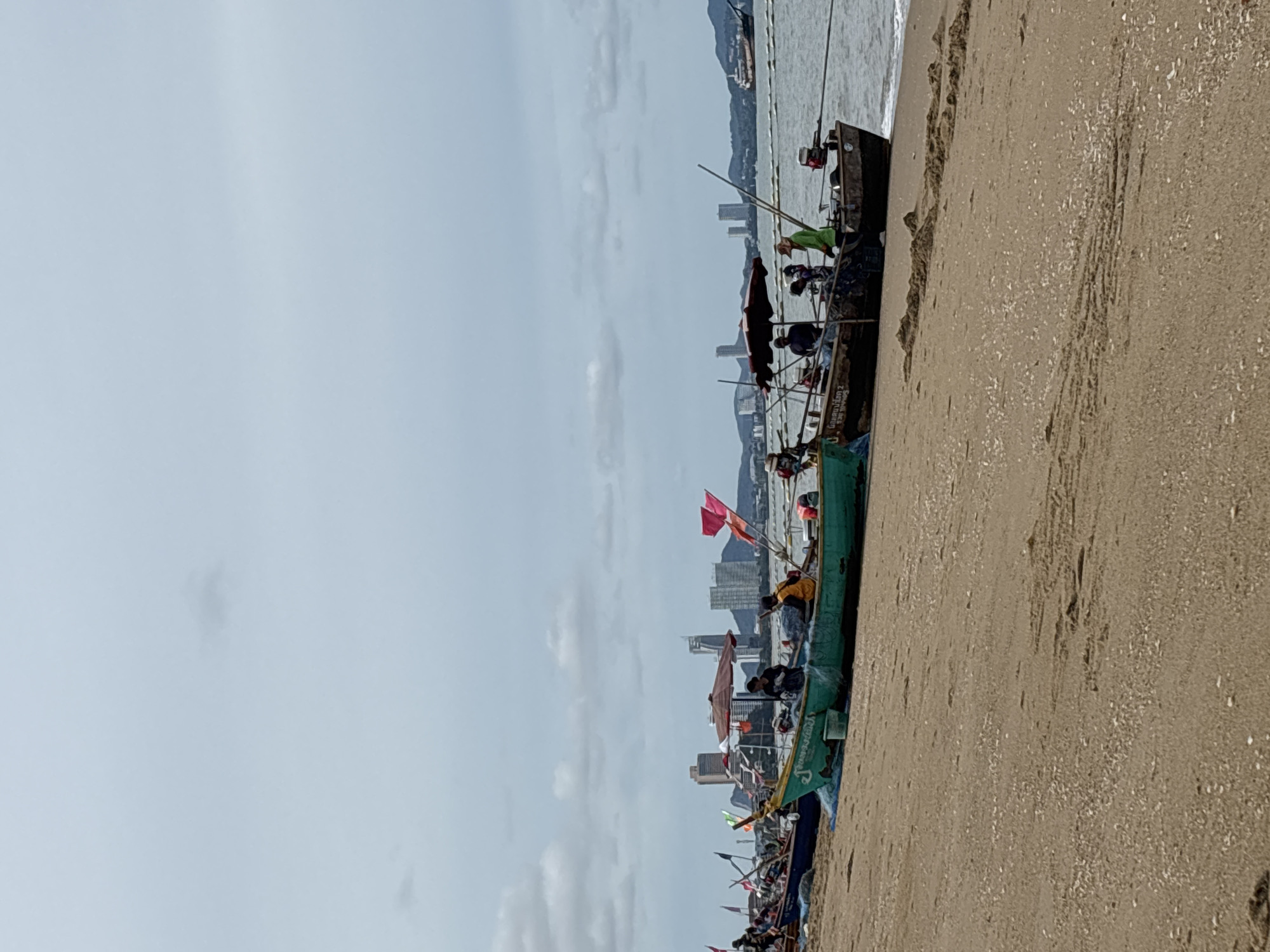 This is a wide shot of a beach in Pattaya City, Thailand. The beach is sandy and appears to be a popular spot for fishing boats. In the foreground, several small, brightly colored fishing boats are pulled up onto the sand. They are equipped with outboard motors and some have umbrellas providing shade for the occupants. Several people are visible in and around the boats, some appearing to be fishermen. The boats are lined up along the shore, with some waves lapping at the waterline. In the background, the cityscape of Pattaya is visible, with numerous high-rise buildings dominating the skyline. The sky is overcast with scattered clouds. The overall scene suggests a typical day for fishing operations in this coastal city.