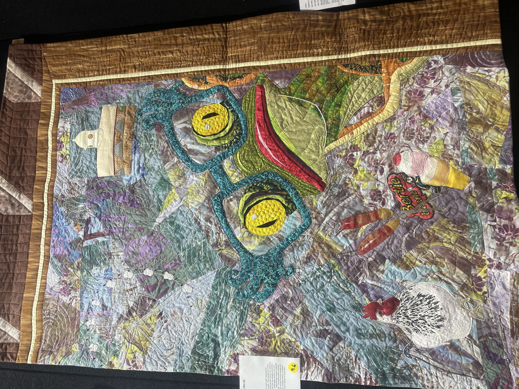 This is a photograph of a quilt displayed indoors. The quilt features a whimsical design with a large, cartoonish frog wearing oversized teal glasses and bright yellow eyes. Below the frog, there is a white chicken with black speckles and a red comb, a pink and yellow ice cream cone, and two red hot dogs on sticks. The background of the quilt is a busy, abstract pattern in shades of green, purple, blue, and brown. The quilt is framed by a brown border with a wood-grain pattern. There is a white label on the left side of the quilt, partially obscured, with text that appears to describe the artwork. Another white label is visible on the right side. The overall impression is of a detailed and colorful textile artwork. The photograph does not contain any information that would allow for the identification of its location within Brisbane, Australia.