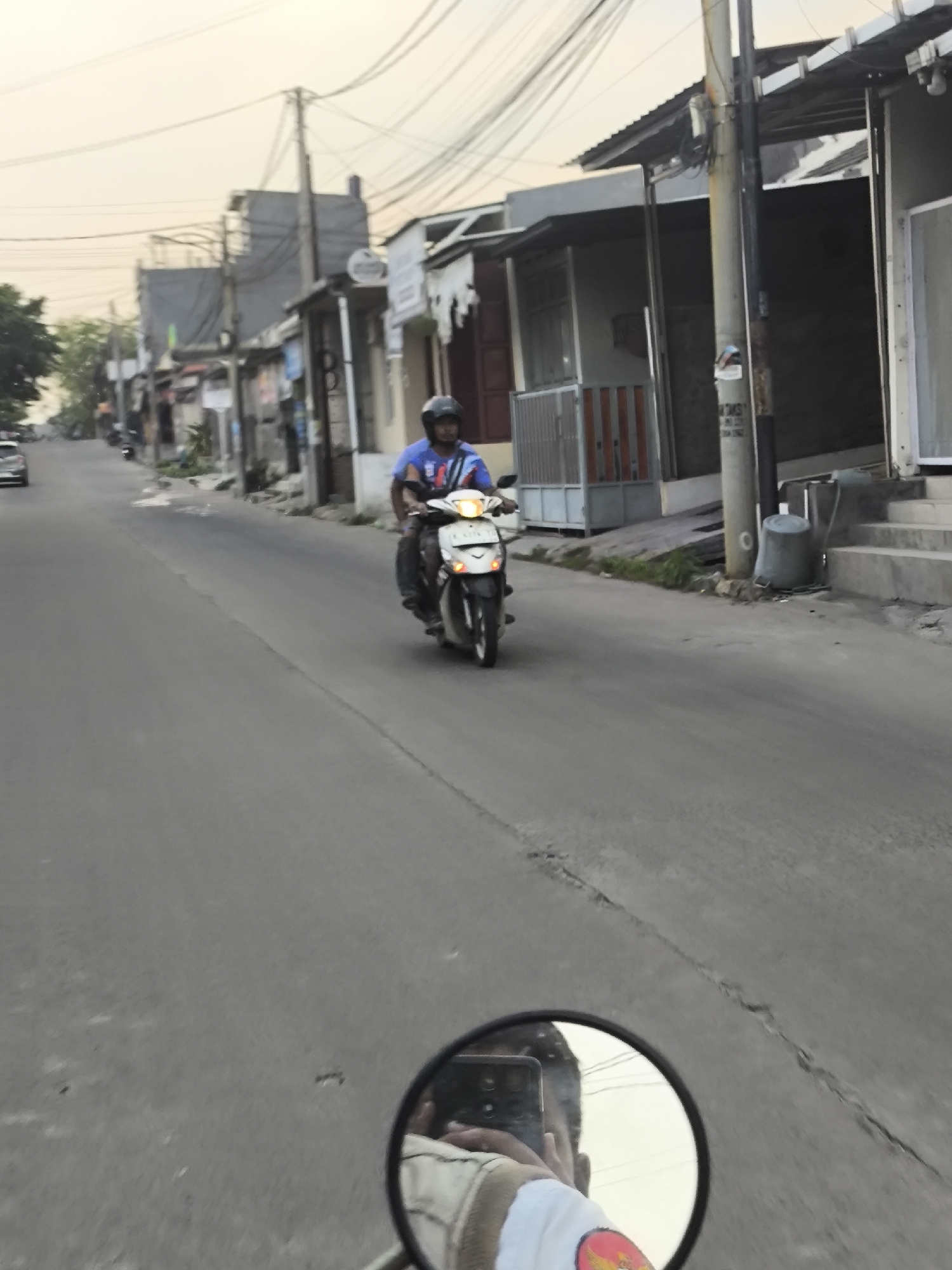 In Cilegon, Indonesia, a person rides a motorcycle down a street lined with buildings and utility poles. The motorcyclist wears a blue shirt and a black helmet, and their face is visible. The motorcycle is white and black with its headlight on. In the foreground, a rearview mirror reflects the motorcyclist's arm holding a phone and their upper body. The street is paved and appears to be slightly uphill. Overhead, numerous electrical wires crisscross the sky. The buildings along the street have various styles, some with awnings and metal fences. The sky is overcast with a pale, yellowish hue, suggesting either early morning or late afternoon light. The scene depicts a common urban streetscape in Indonesia.