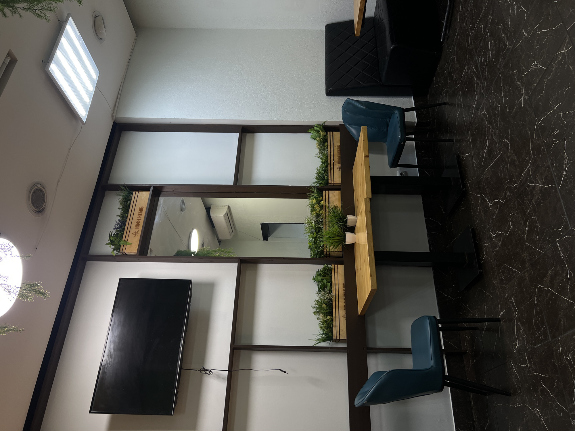 The image depicts the interior of a cafe in Saint Petersburg, Russia, featuring a modern and clean aesthetic. In the foreground, two teal chairs with black metal legs are positioned around a light-colored wooden table with a black pedestal base. The table has a dark marble-patterned tile floor beneath it.
To the right of the table, a black quilted bench seat is visible, also with a dark marble-patterned floor. Another light-colored wooden table, partially visible on the right edge of the image, suggests further seating arrangements.
The background reveals a wall-mounted black television screen on the left. Behind the table and chairs, a decorative wooden structure with shelves holds several wooden planters filled with green plants. This structure is set against a wall with framed sections. A mirror is incorporated into this wall feature, reflecting the room. An air conditioning unit is visible on the wall in the reflected area.
Above, a rectangular fluorescent light fixture illuminates the space, and decorative greenery hangs from the ceiling near a circular light fixture. The overall impression is one of a contemporary and comfortable dining or waiting area.