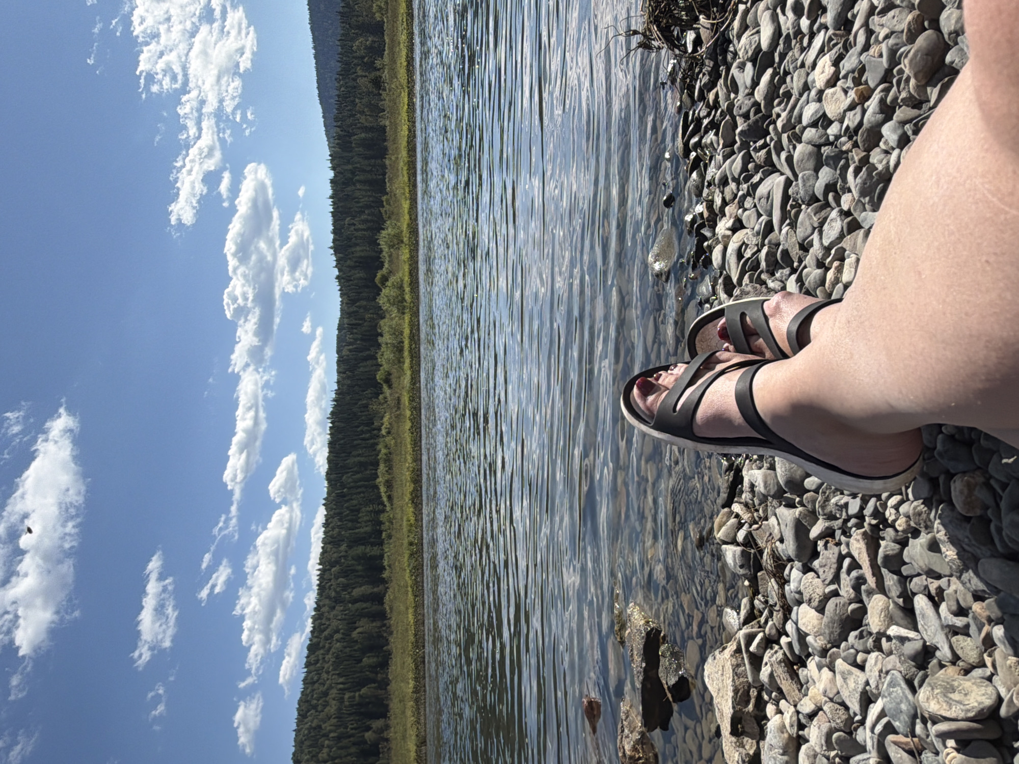 A person's legs, wearing black sandals, are extended towards the viewer, resting on a rocky shoreline. The shoreline is composed of various sizes of grey, brown, and white stones. To the left, the clear water of a river is visible, with the riverbed's stones discernible beneath the surface. The river flows horizontally across the image, reflecting the blue sky and white clouds. Beyond the river, a dense forest of green trees covers rolling hills, stretching towards the horizon under a bright, cloudy sky. The location is Ust-Kabirza, Russia.