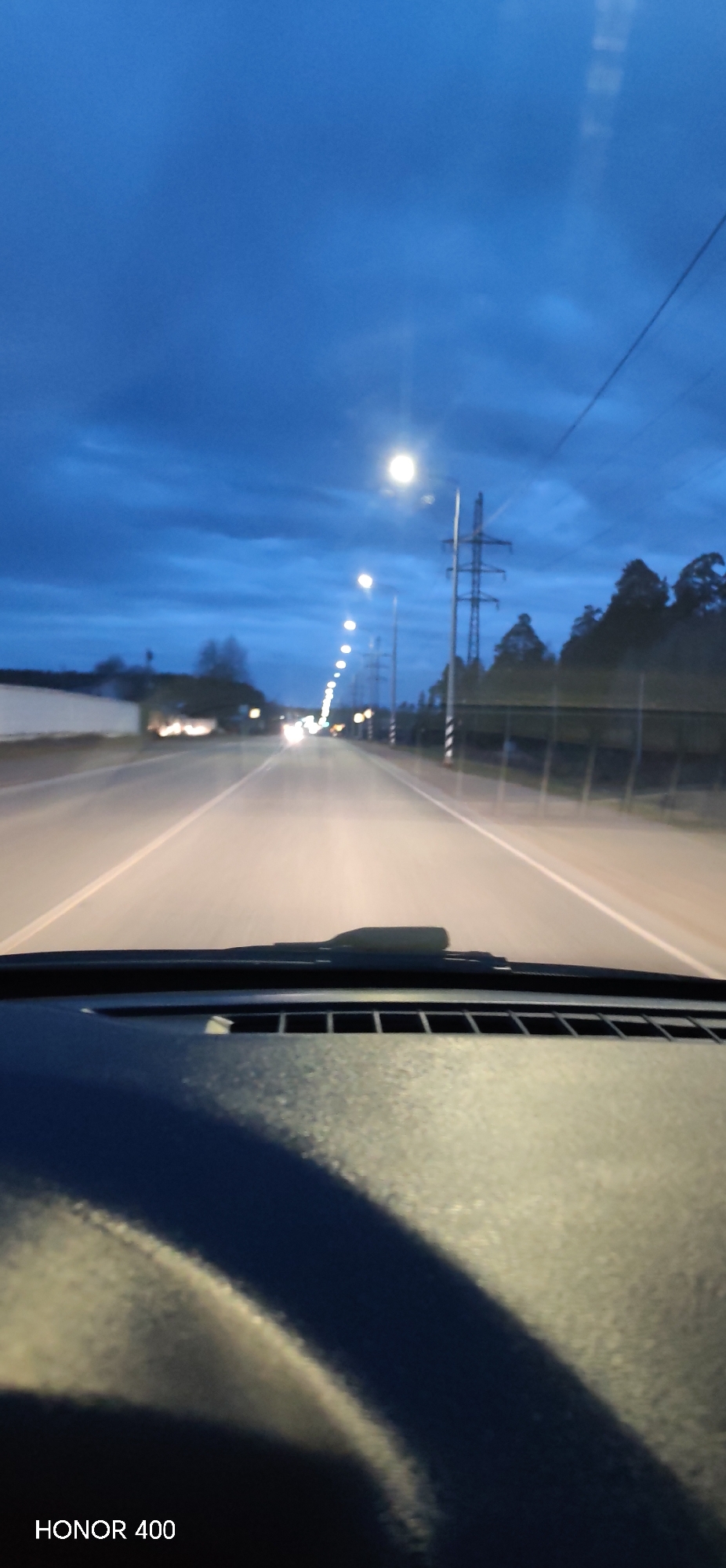 The image depicts a view from inside a car driving on a road. The scene is dominated by the road ahead, illuminated by streetlights. There is a row of streetlights lining the road, which extends into the distance. Power lines are visible alongside the road, supported by utility poles. A treeline can be seen on the right side of the road. The sky is overcast, with dark blue clouds filling the upper portion of the image. The dashboard and steering wheel of the car are in the foreground, creating a first-person perspective of driving. The text "HONOR 400" is visible in the bottom left corner. The location is likely Chaikovsky, Russia, based on the streetlights and infrastructure.