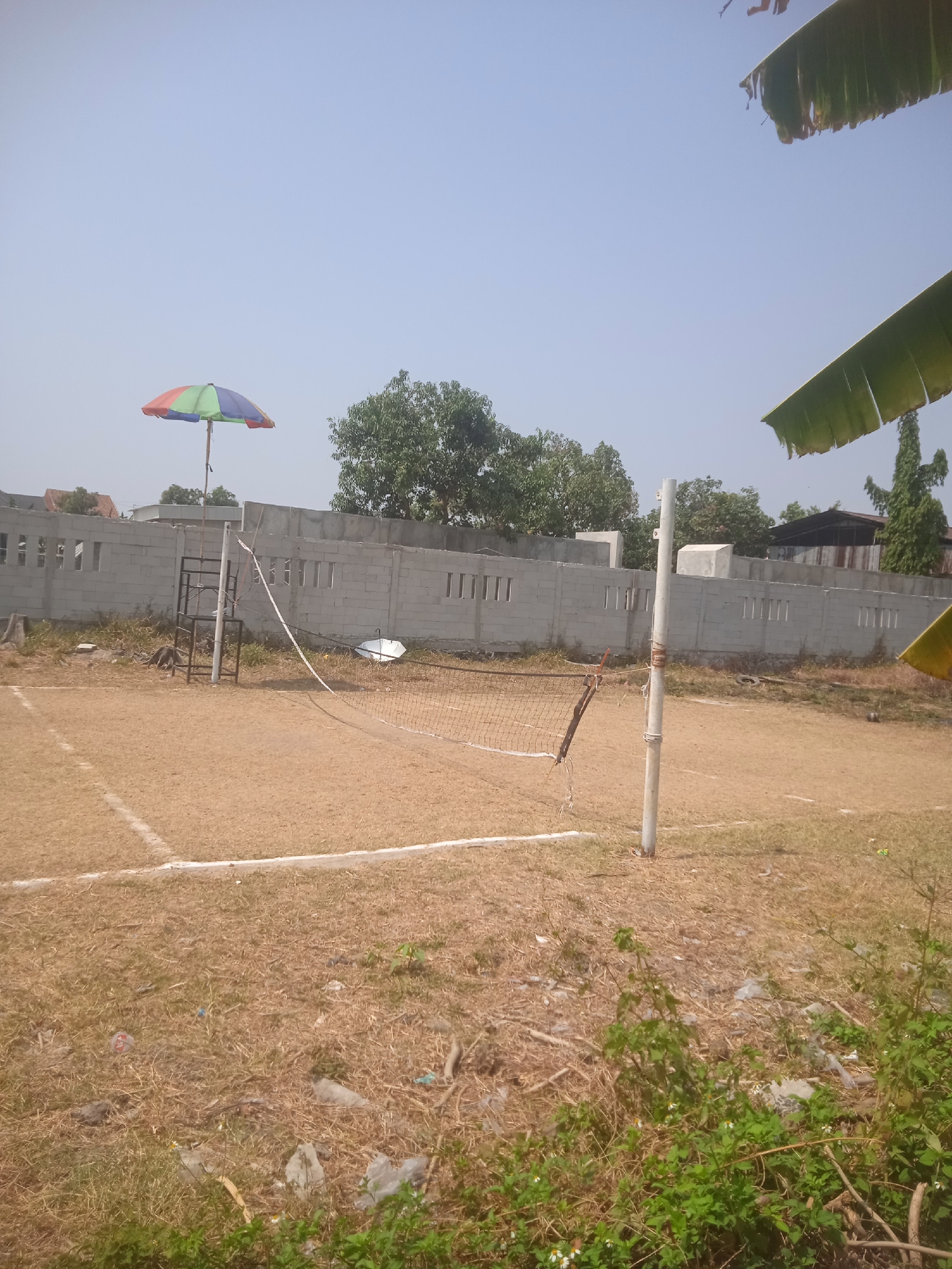 The image depicts an outdoor volleyball court with a sandy surface, marked by white lines. A net is set up across the court, supported by poles. A colorful umbrella is positioned near one of the net posts, attached to a metal stand. In the background, a low brick wall with rectangular openings stretches across the scene. Beyond the wall, trees and buildings are visible under a clear sky. The court area is littered with dry grass and some scattered trash, suggesting it may be in a less-maintained condition or an area adjacent to a more populated zone. The presence of the umbrella and the court suggests it's a recreational area, possibly within or near Klodran, Indonesia.