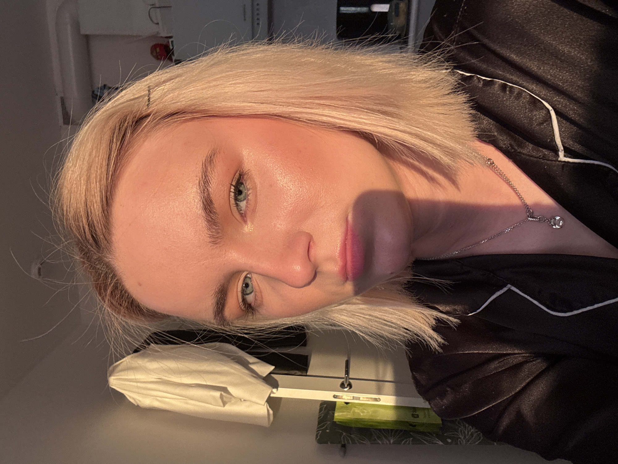 A close-up, rotated headshot of a fair-skinned woman with short, blonde hair and light blue eyes. She is wearing a dark, satin pajama top with white piping and a delicate silver necklace. The background includes a white surface with a folded white cloth, a white electrical outlet panel, and a green object with a patterned edge. The photo appears to be taken indoors, likely in a residential setting. There is nothing in the image to indicate the city of Vsevolozhsk, Russia.
