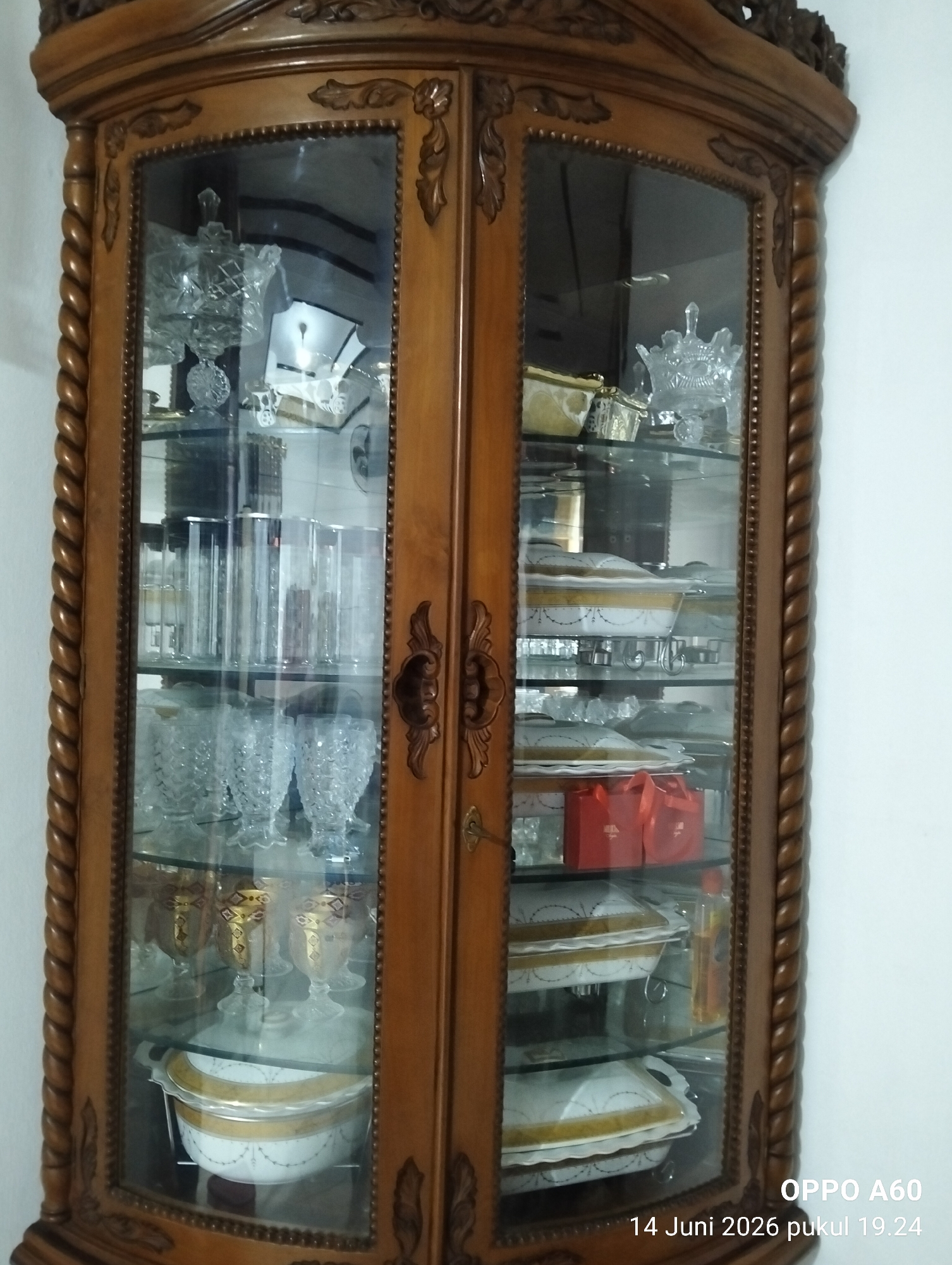 The image shows an antique wooden display cabinet filled with various tableware. The cabinet has a carved wooden frame and glass doors, revealing multiple shelves stocked with items. On the top shelf, there are clear glass decorative pieces. Below, there are rows of tall, clear glasses. Further down, a collection of decorative crystal bowls is visible. The middle shelves display serving dishes with white and gold ornate patterns, some with lids. There are also two red gift bags on one of the lower shelves. The bottom shelves contain more serving dishes and a decorative glass vase with amber liquid. The cabinet is positioned against a plain white wall. The bottom right corner of the image contains a timestamp that reads "OPPO A60 14 Juni 2026 pukul 19.24". The location is stated as Payagaboh, Indonesia.