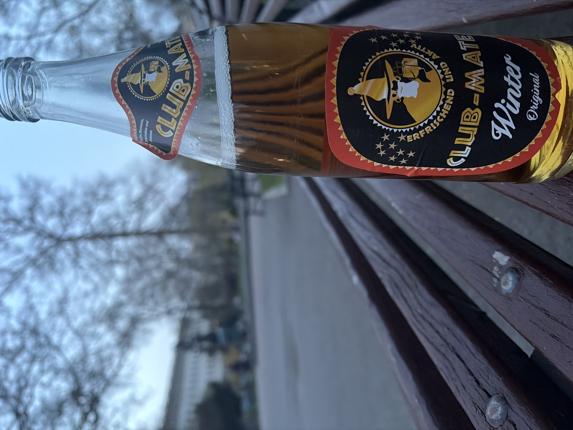 Here is a concise description of the image:
The image shows a bottle of Club-Mate Winter Original on a wooden bench. The label is visible, with "Club-Mate Winter" in large letters and "Original" underneath. The label also includes the "Club-Mate" logo and the text "ERFRISCHEND UND AKTIV". The background shows trees and buildings out of focus. The exact location in Sofia, Bulgaria, is impossible to determine from the image without further information. There are no people visible in the image.