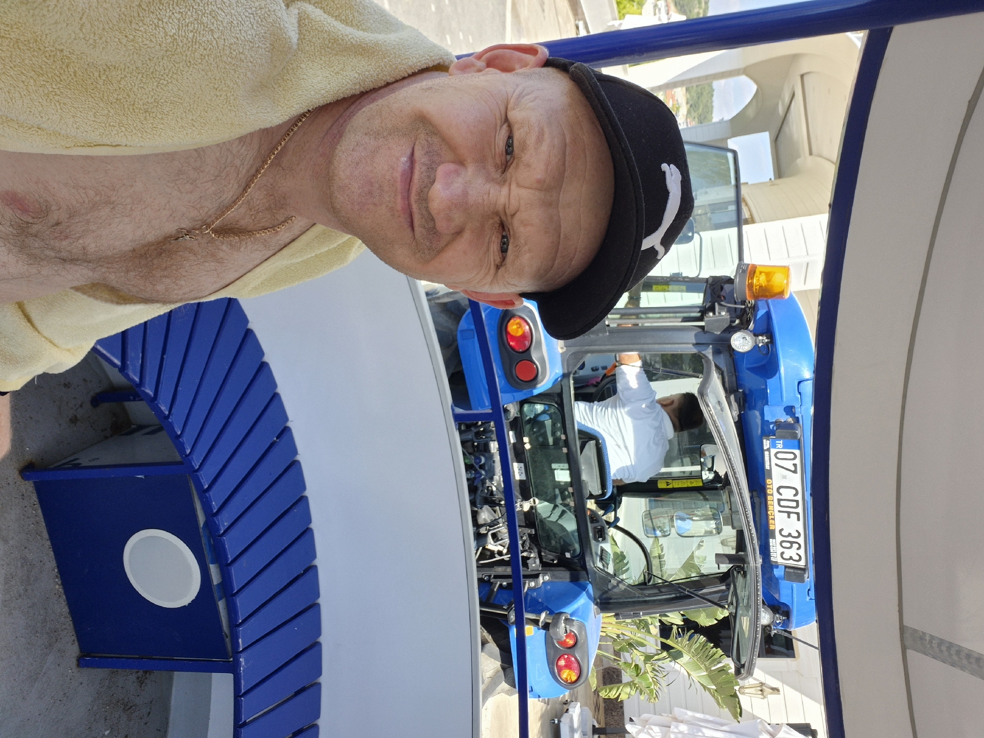 The image is a selfie taken outdoors. A middle-aged man with fair skin, wearing a black Puma baseball cap and a yellow towel draped over his shoulder, is the primary subject. He has a gold necklace visible on his chest.
Behind him, a blue tractor is visible with a person in the driver's seat wearing a white shirt. The tractor's license plate reads "07 CDF 363". To the left of the man, there is a curved blue bench. The background shows a blurred landscape, including buildings and vegetation.
Based on the license plate and the overall environment, the location is likely Alanya, Turkey. The presence of the tractor suggests the photo was taken in a semi-urban or rural area within Alanya.