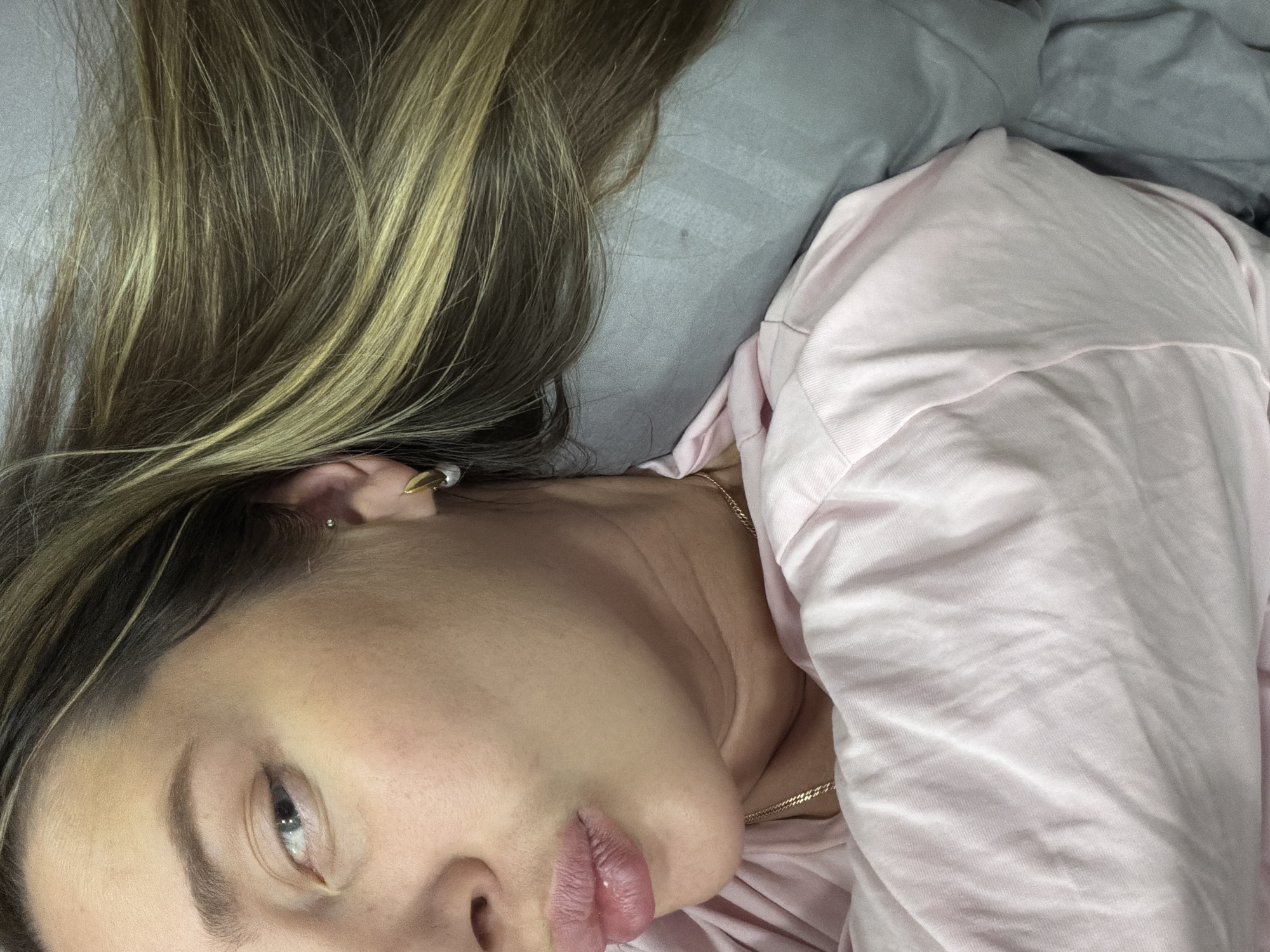 A woman with fair skin and light brown hair with blonde highlights is lying down. She is wearing a pink garment and resting her head on a light gray pillow. She has a gold-colored earring and a gold-colored necklace. The background is out of focus. The location is not discernible from the image.