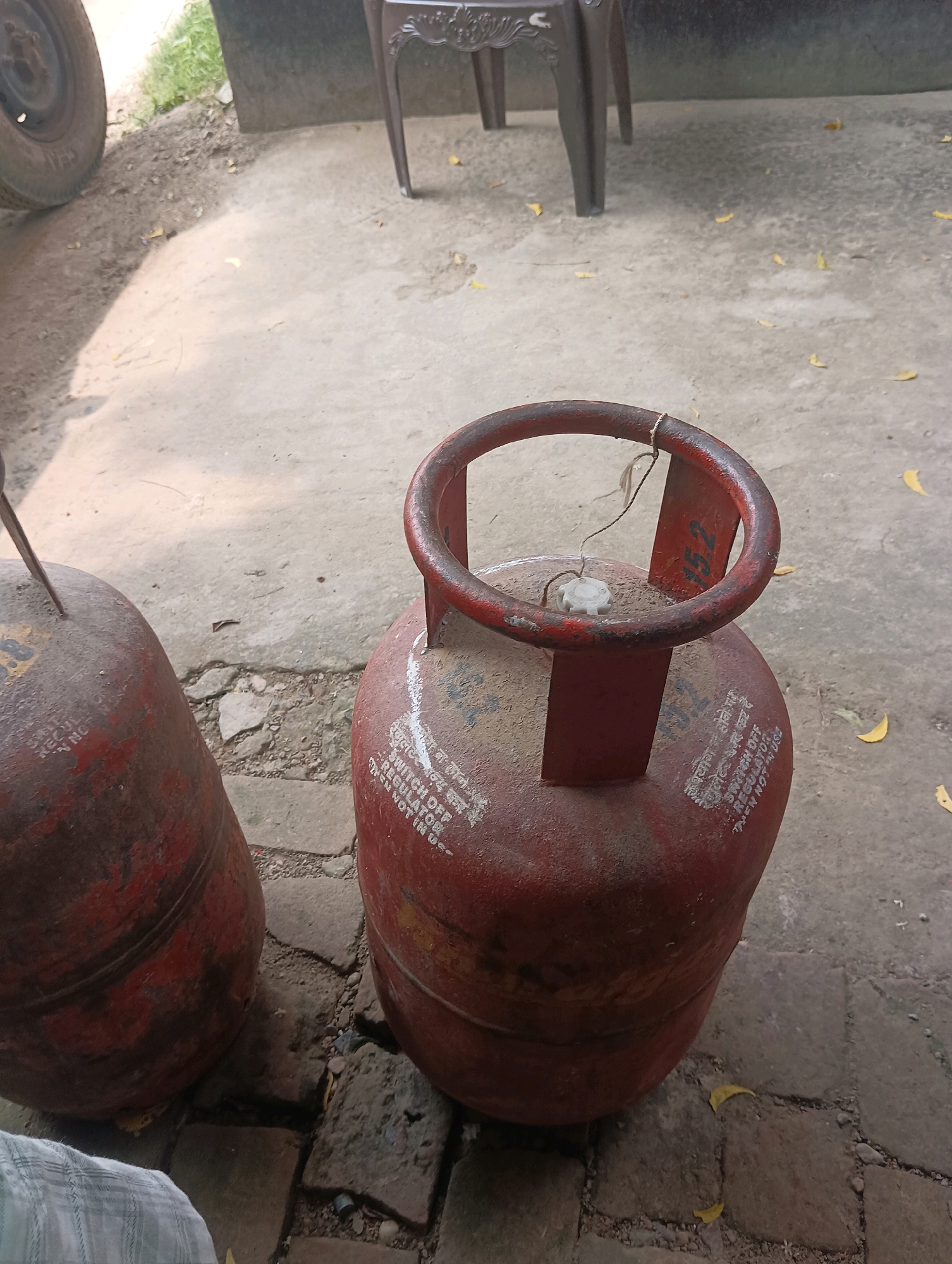 In an outdoor, ground-level, medium-shot image, two red, metal LPG cylinders are positioned on a concrete and brick surface. The cylinder in the foreground is upright and faces the viewer at an angle, showing its top valve and handle. The second cylinder is to the left, also red and metal, tilted on its side, with its valve cap missing. Both cylinders have markings on them; the foreground cylinder has white text that appears to be safety instructions in Hindi and English. The background includes a dark brown, ornate plastic chair, the side of a dark-colored vehicle with a visible tire, and sparse green vegetation. The ground surface is a mix of concrete slabs and rough bricks, with scattered dry leaves. The lighting suggests it is daytime, with shadows visible on the ground.