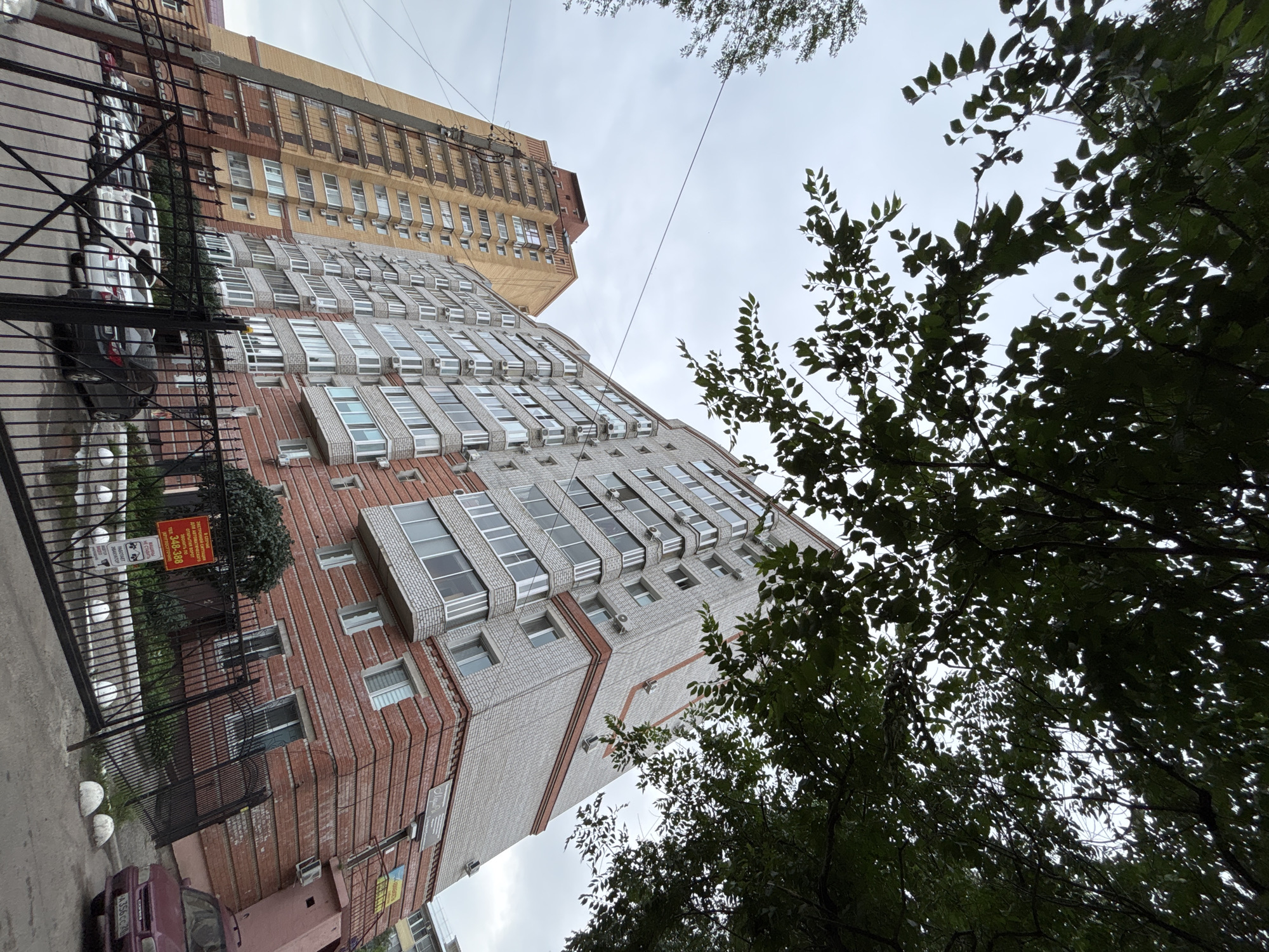 The image shows an exterior view of two apartment buildings, taken from a low angle, looking upwards. The buildings are modern and multi-story, with visible architectural details like balconies and air conditioning units. The building in the foreground is clad in a combination of light-colored brick and darker red brick, with many windows. The building behind it is taller and appears to be older, with a yellowish-brown facade and rows of windows. Trees with dense green foliage are in the foreground and partially obscure the buildings, suggesting a natural setting within an urban environment. The sky is overcast and gray, indicating cloudy weather. The scene appears to be a street or courtyard area in front of the buildings, with a black metal fence and some parked cars visible. There is a red sign on the fence with Russian text. The overall impression is one of a residential area on a possibly cool or overcast day.
