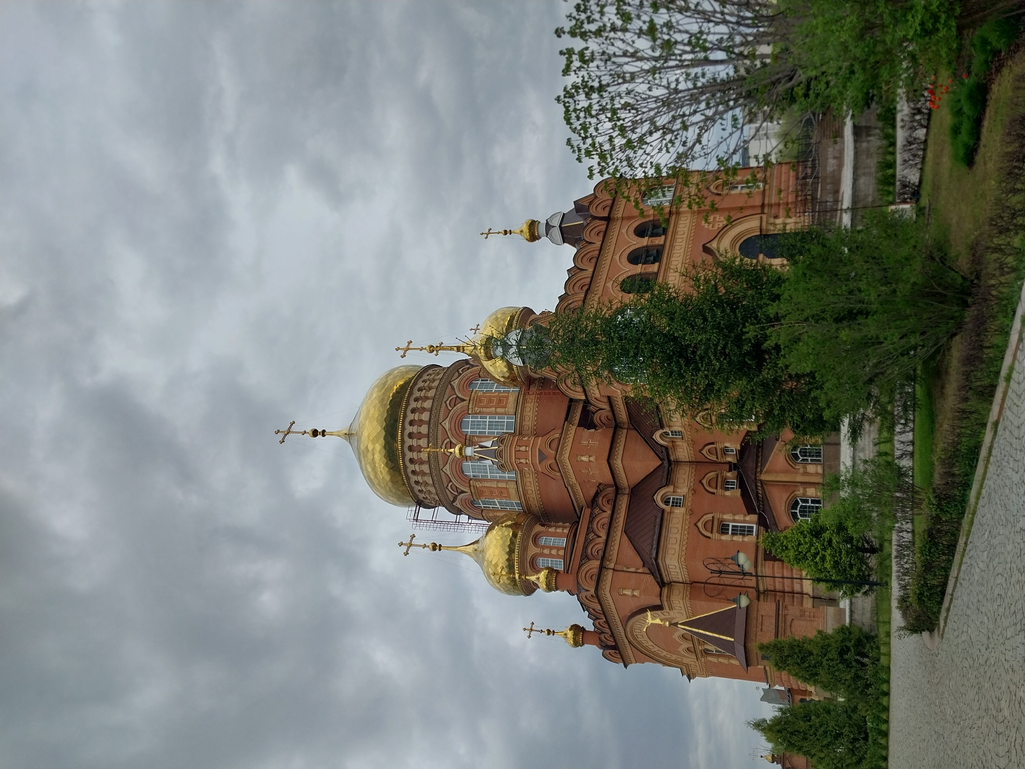 This image shows the Orenburg Eparchial Theological Seminary located in Orenburg, Russia. The image is captured from a low angle, emphasizing the building's height and golden domes. The building is constructed of red brick and has several ornate golden domes topped with crosses. The sky is overcast. The foreground shows greenery and trees. The perspective of the photograph appears to be rotated ninety degrees counter-clockwise.