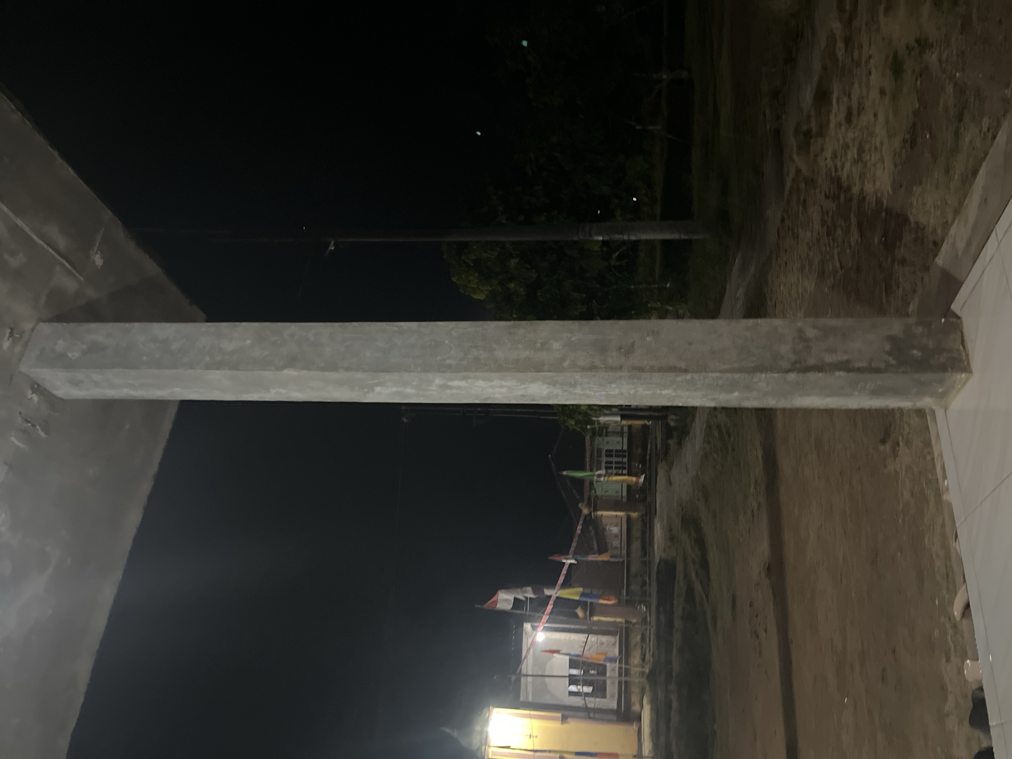 The image captures an outdoor scene at night, possibly in Kebumen, Indonesia, as suggested by the flags which appear to be the Indonesian flag. In the foreground, a large, rough-textured concrete pillar dominates the right side of the frame, partially obscuring the view. A concrete overhang extends from the top of the frame.
In the background, a building with illuminated windows is visible, along with several flags on poles. One prominent flag, red and white, is mounted on a pole. Other smaller, colorful flags are also present. A street lamp or similar pole is situated to the right of the building. The ground appears to be unpaved, with areas of dirt and sparse vegetation. Trees and foliage are visible further in the background, silhouetted against the dark night sky. The overall lighting suggests artificial illumination from the building and the street lamp, casting shadows and creating a dimly lit atmosphere. There are no people visible in the image.