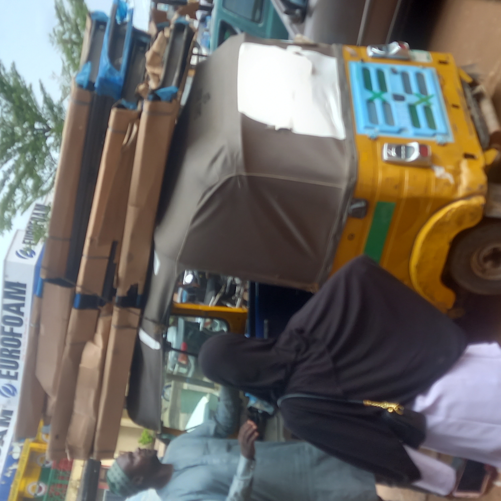 A yellow motorized tricycle (auto-rickshaw) is heavily loaded with stacked rectangular packages marked "EUROFOAM." The vehicle is parked on a city street in Sardauna, Nigeria. An adult male, wearing a white shirt and cap, stands beside the auto-rickshaw. Another individual, draped in a black hijab, is positioned next to the auto-rickshaw. The scene suggests a commercial transaction involving the transportation of goods.