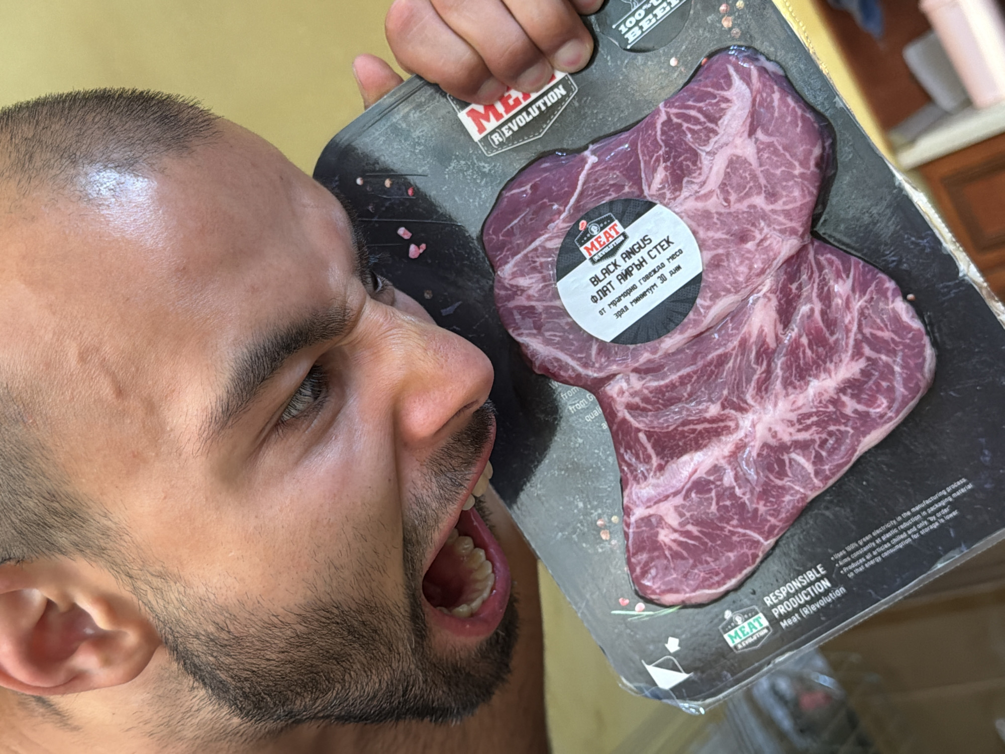A close-up photograph captures a man humorously posing with a vacuum-sealed package of raw beef steak held near his wide-open mouth, as if expressing an intense desire to eat it. The man, with short dark hair, stubble, and an expressive left eye, looks directly at the packaged meat. His mouth is open, revealing his teeth and tongue in an exaggerated gesture. He uses his right hand to hold the black packaging, which displays two richly marbled cuts of beef.
The visible text on the packaging prominently features the brand "MEAT REVOLUTION" and declares "100% BEEF". A circular label identifies the product as "BLACK ANGUS", "ФЛАТ АЙРЪН СТЕК" (Flat Iron Steak), "от мраморно говеждо месо" (from marbled beef), and states it is "зрял минимум 30 дни" (aged minimum 30 days). Additional text promotes "RESPONSIBLE PRODUCTION", mentioning the use of "100% green electricity", efforts towards "plastic reduction", production of "chilled and only 'by order'", and lower "energy consumption for storage". The background, out of focus, shows a light-colored wall and some dark furniture, suggesting an indoor setting. This comedic scene takes place potentially in Burgas, Bulgaria.